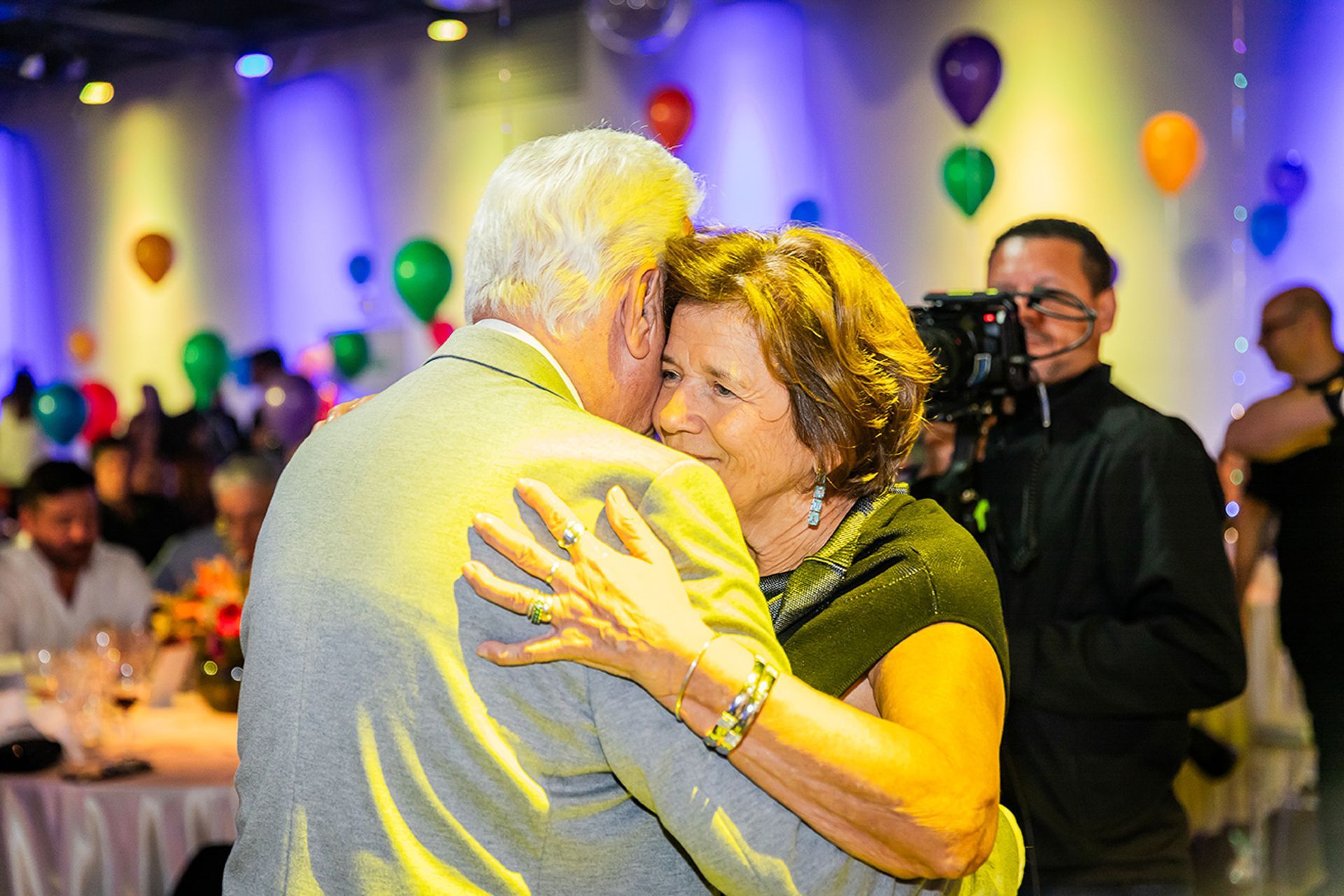 Duas pessoas se abraçam em uma festa decorada com balões, em um espaço festivo com iluminação suave.