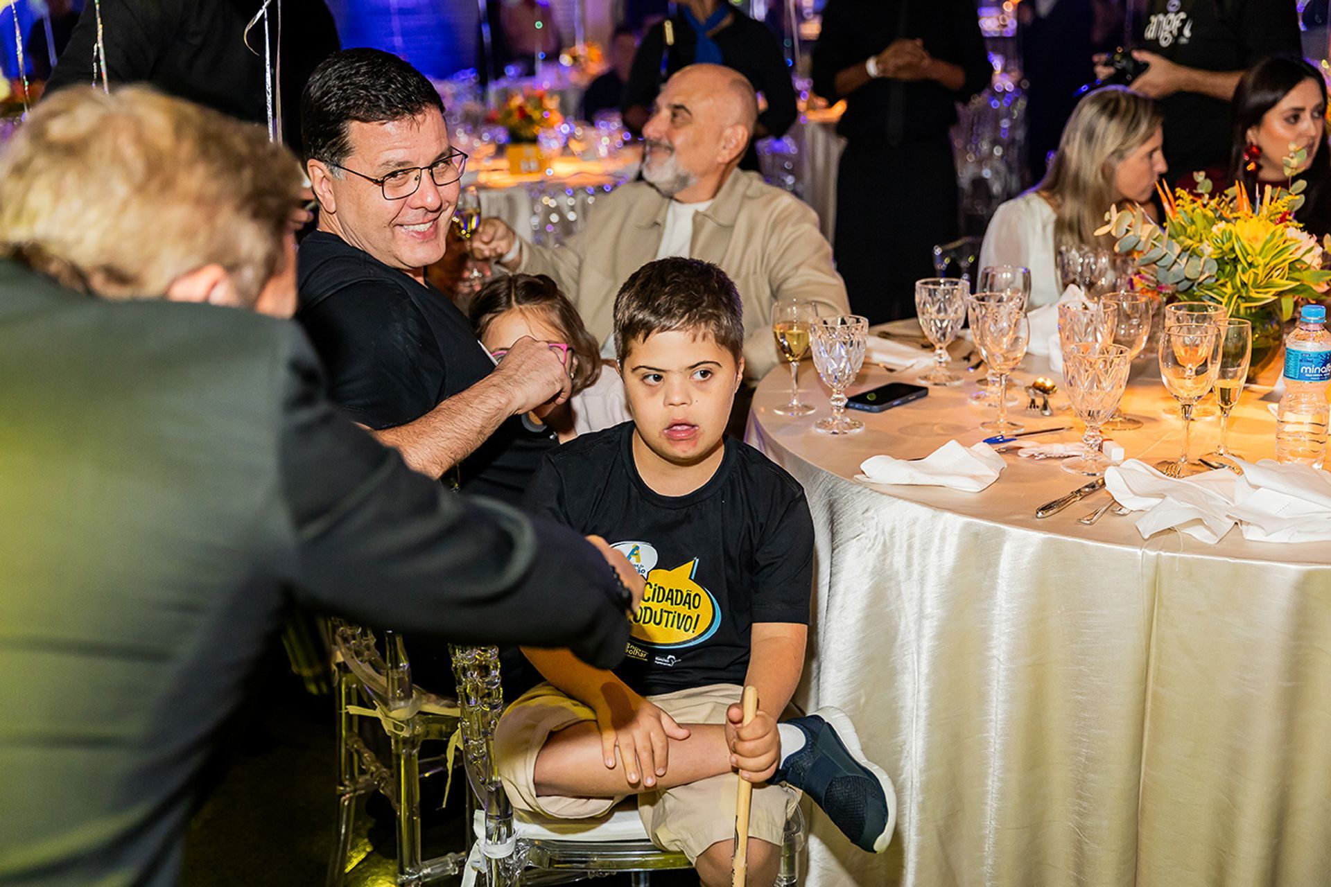 Uma criança está sentada no colo de um adulto em uma mesa de banquete formal com outros convidados.