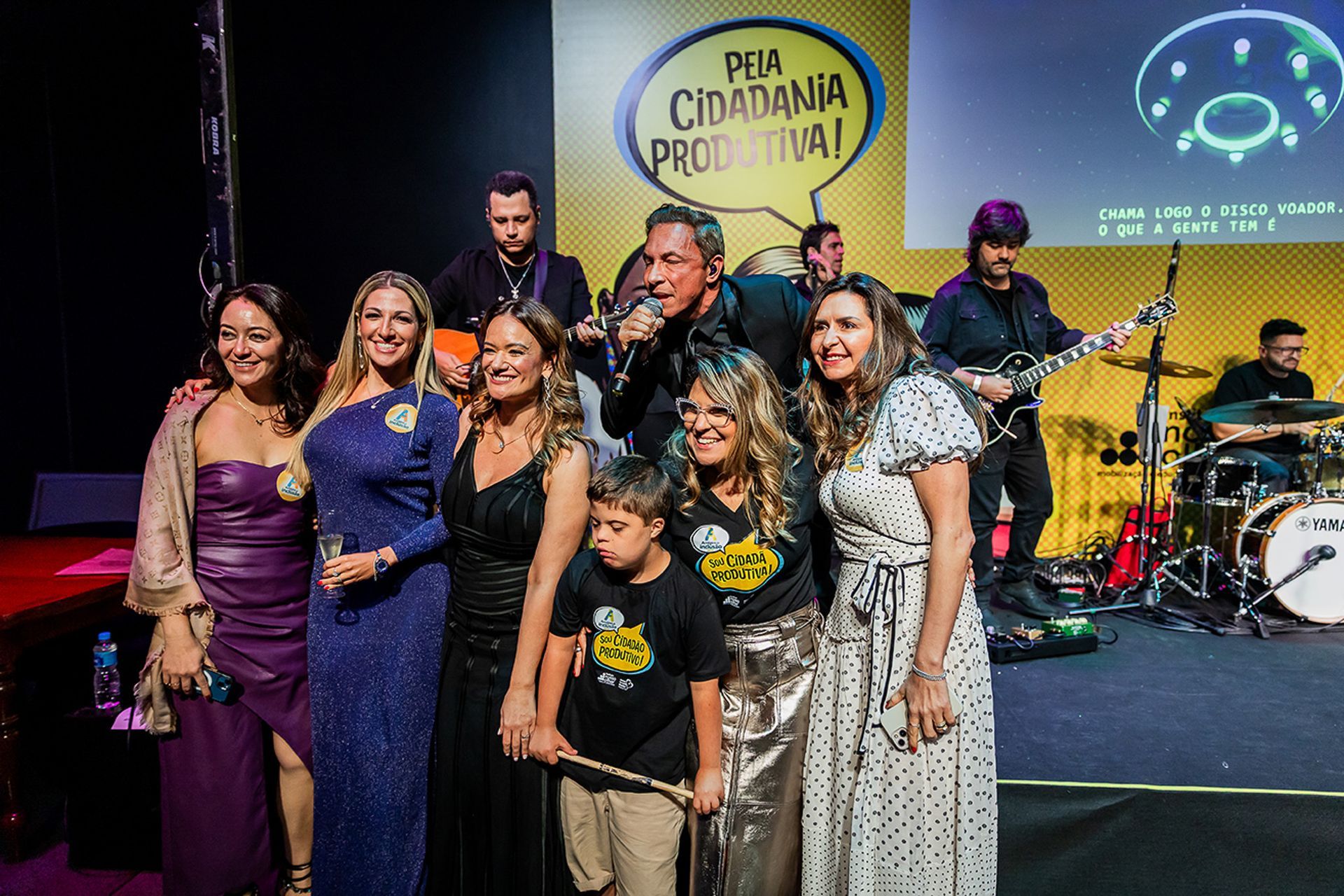 Um grupo de pessoas em pé em um palco durante um evento, sob uma placa amarela com os dizeres 