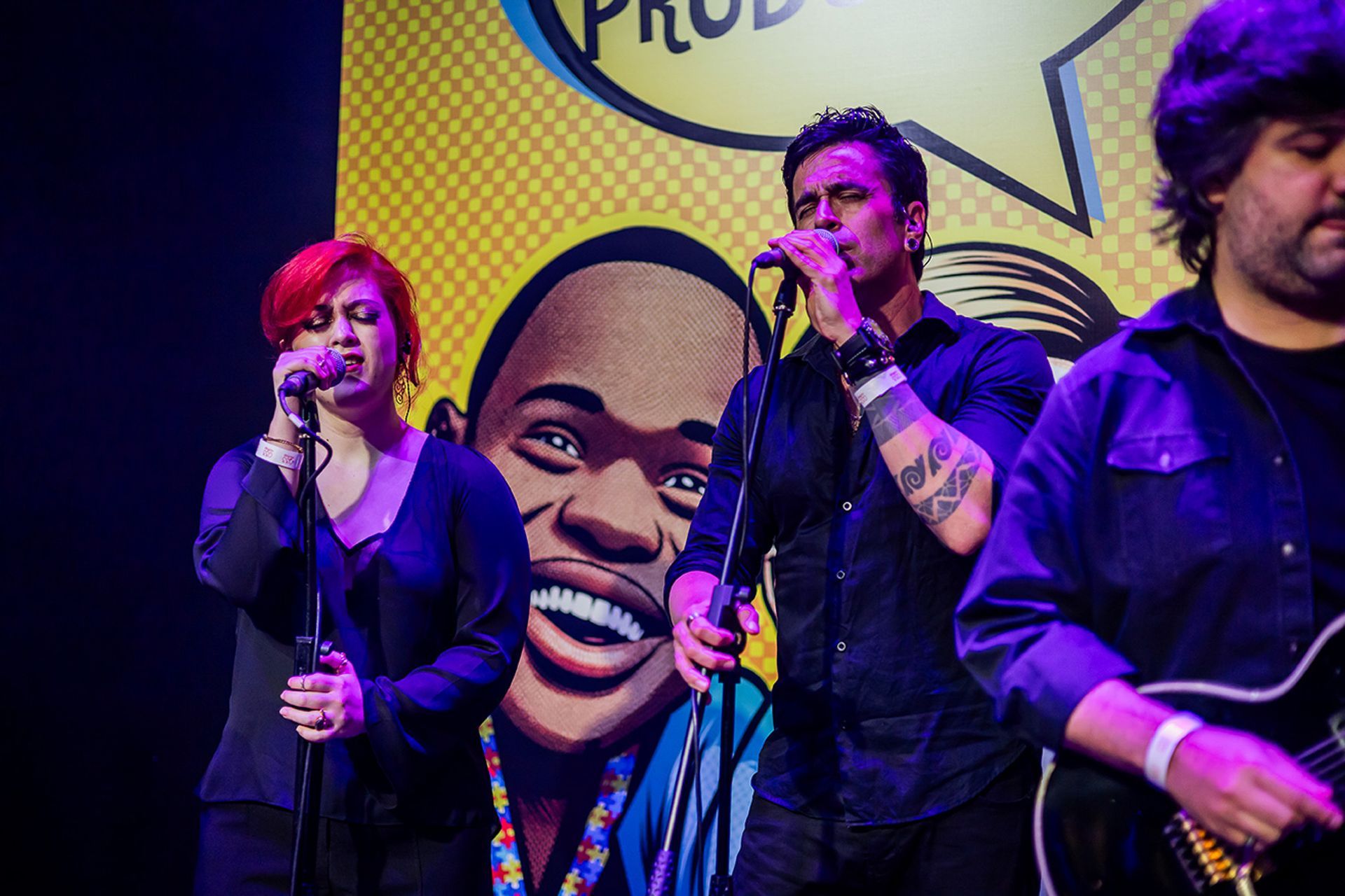 Uma cantor e um cantor se apresentam no palco em frente a um cenário com um rosto de desenho animado colorido e ilustrado.