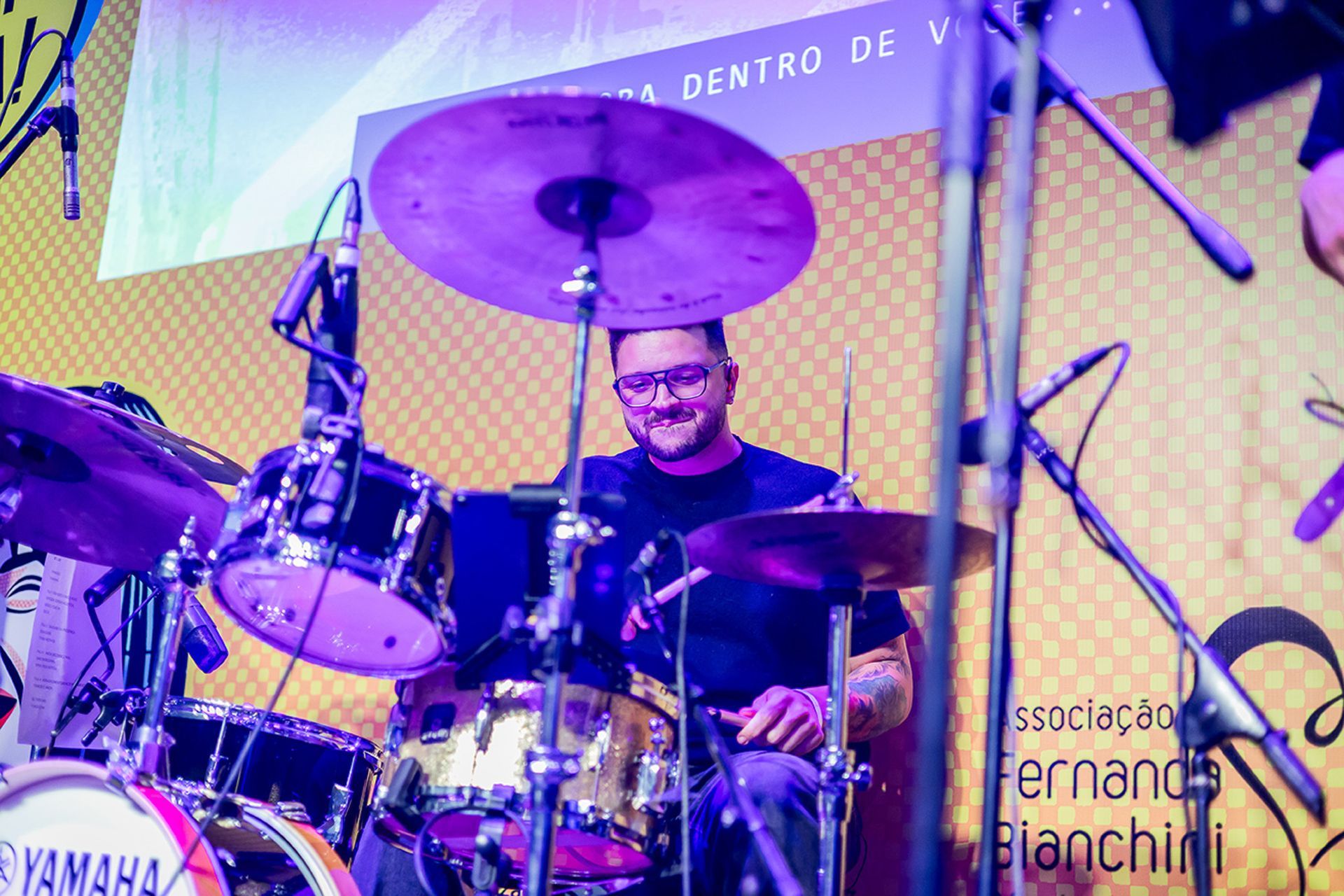 Um baterista sorridente está sentado em uma bateria Yamaha no palco, iluminado por luzes roxas contra um fundo amarelo.