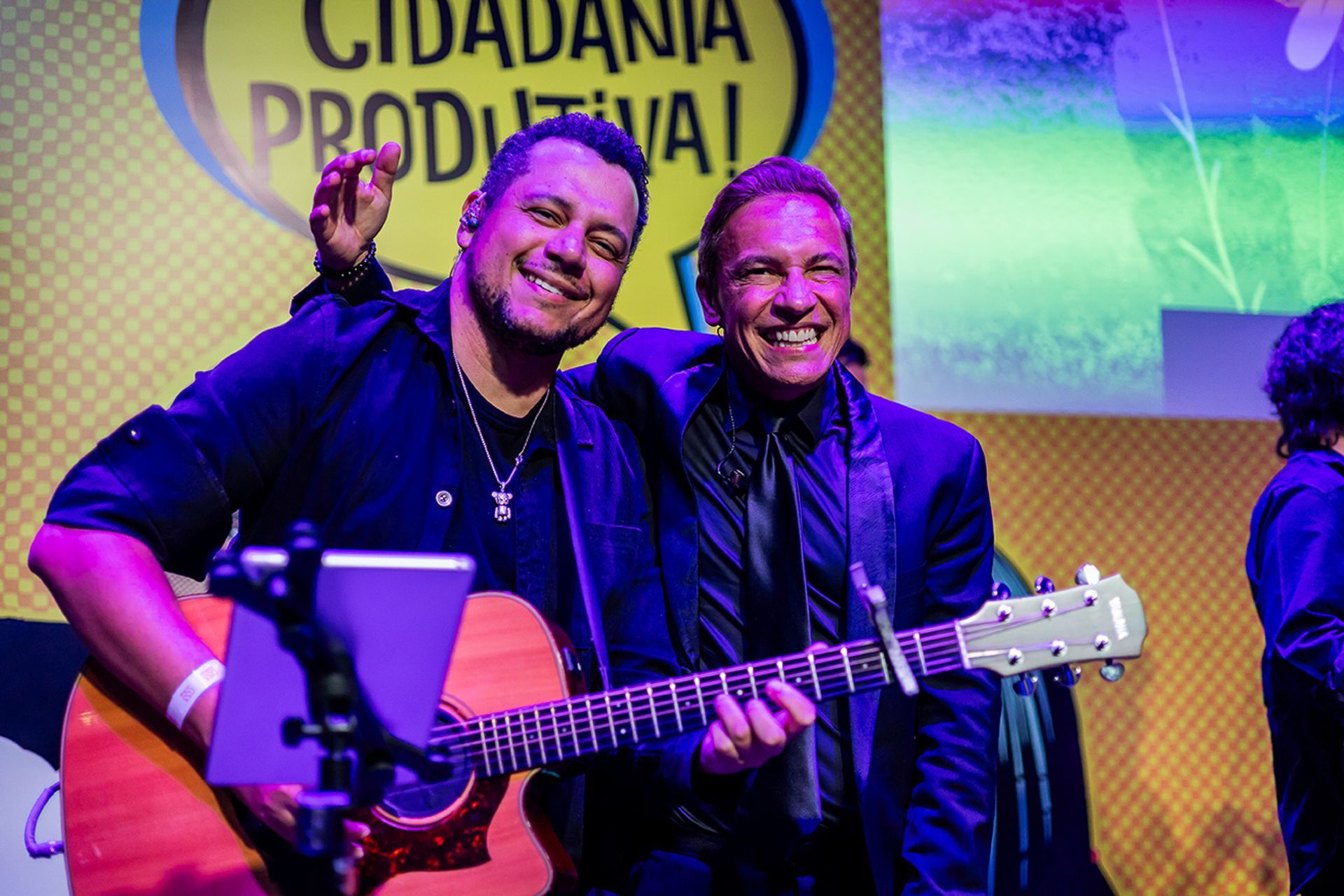Dois artistas sorrindo no palco; um toca violão, enquanto o outro o abraça.