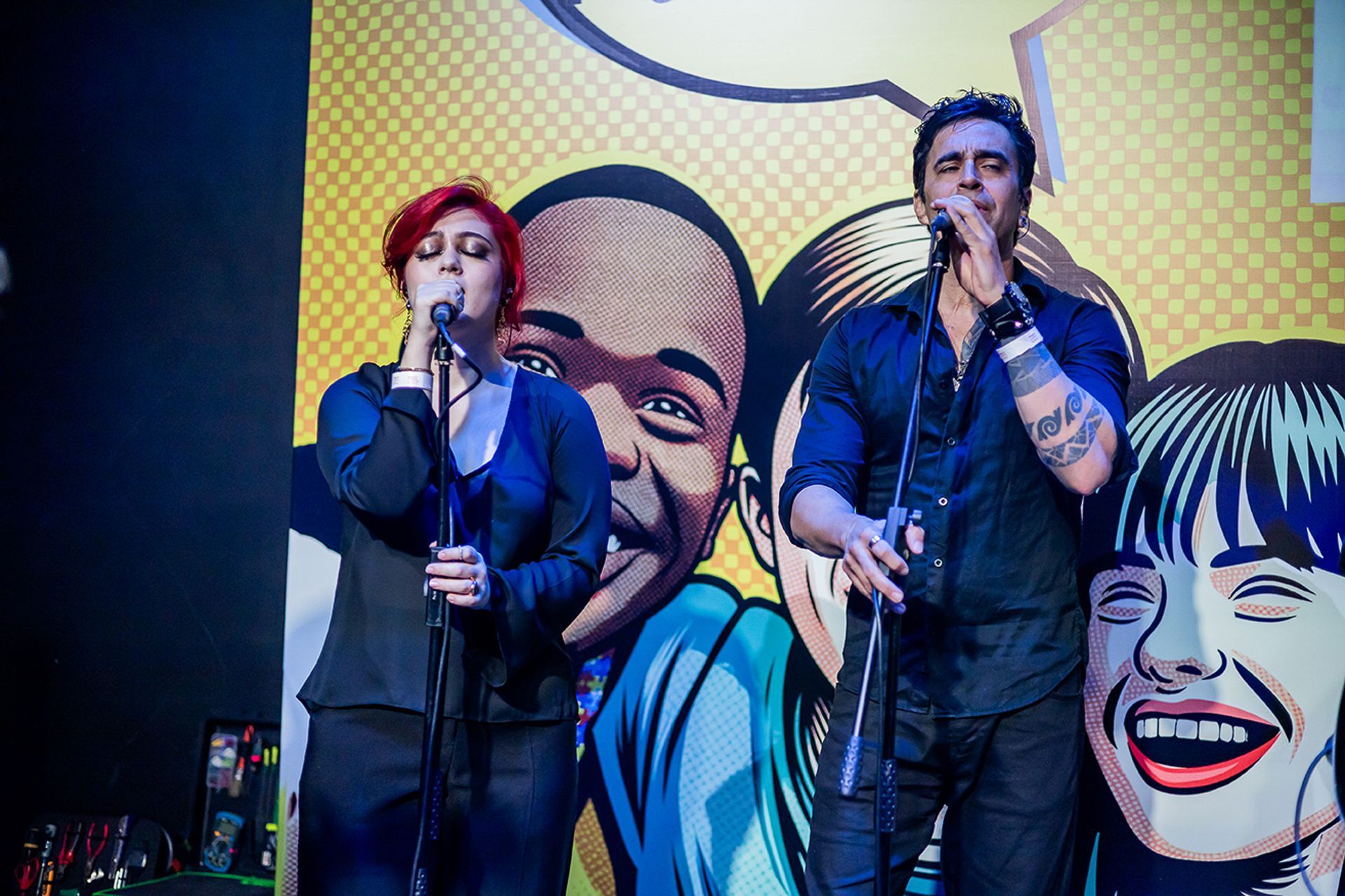 Uma pessoa com cabelo curto e ruivo e uma pessoa com cabelo escuro cantam em frente a um mural em estilo de quadrinhos.