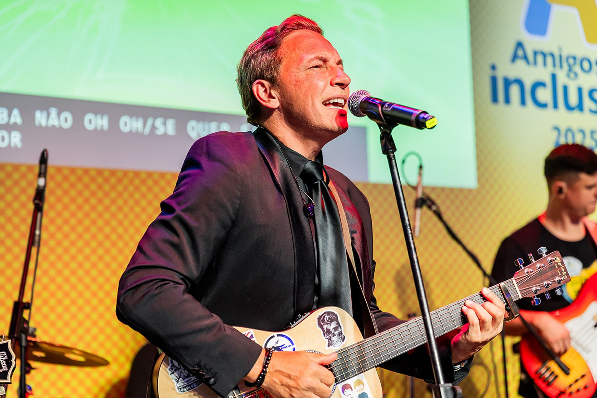 Uma pessoa cantando ao microfone e tocando violão no palco durante um evento.