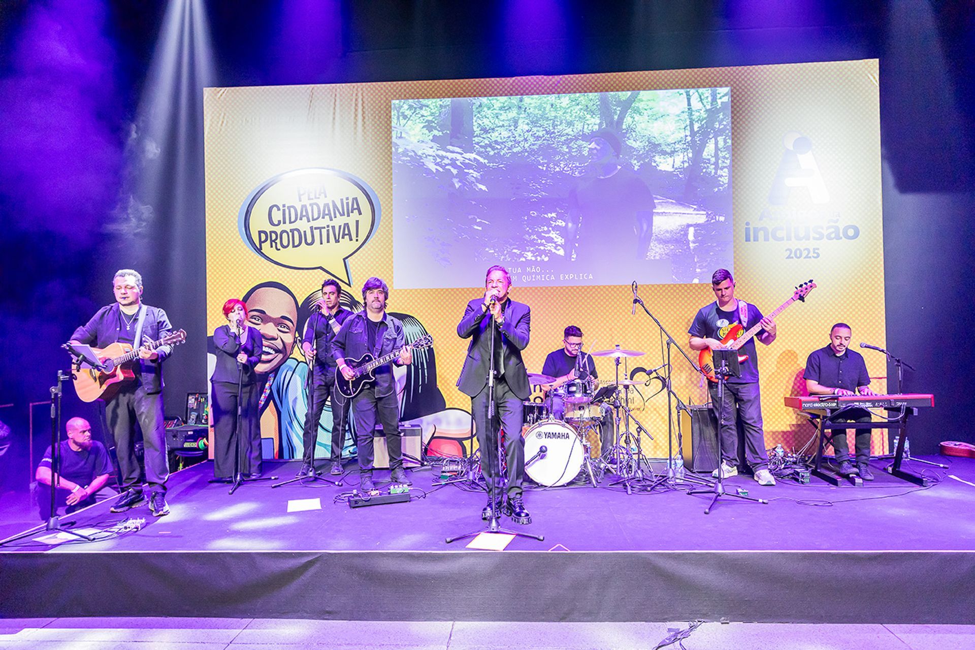 Uma banda se apresenta em um palco em frente a um fundo amarelo com uma tela de projeção durante um evento ao vivo.