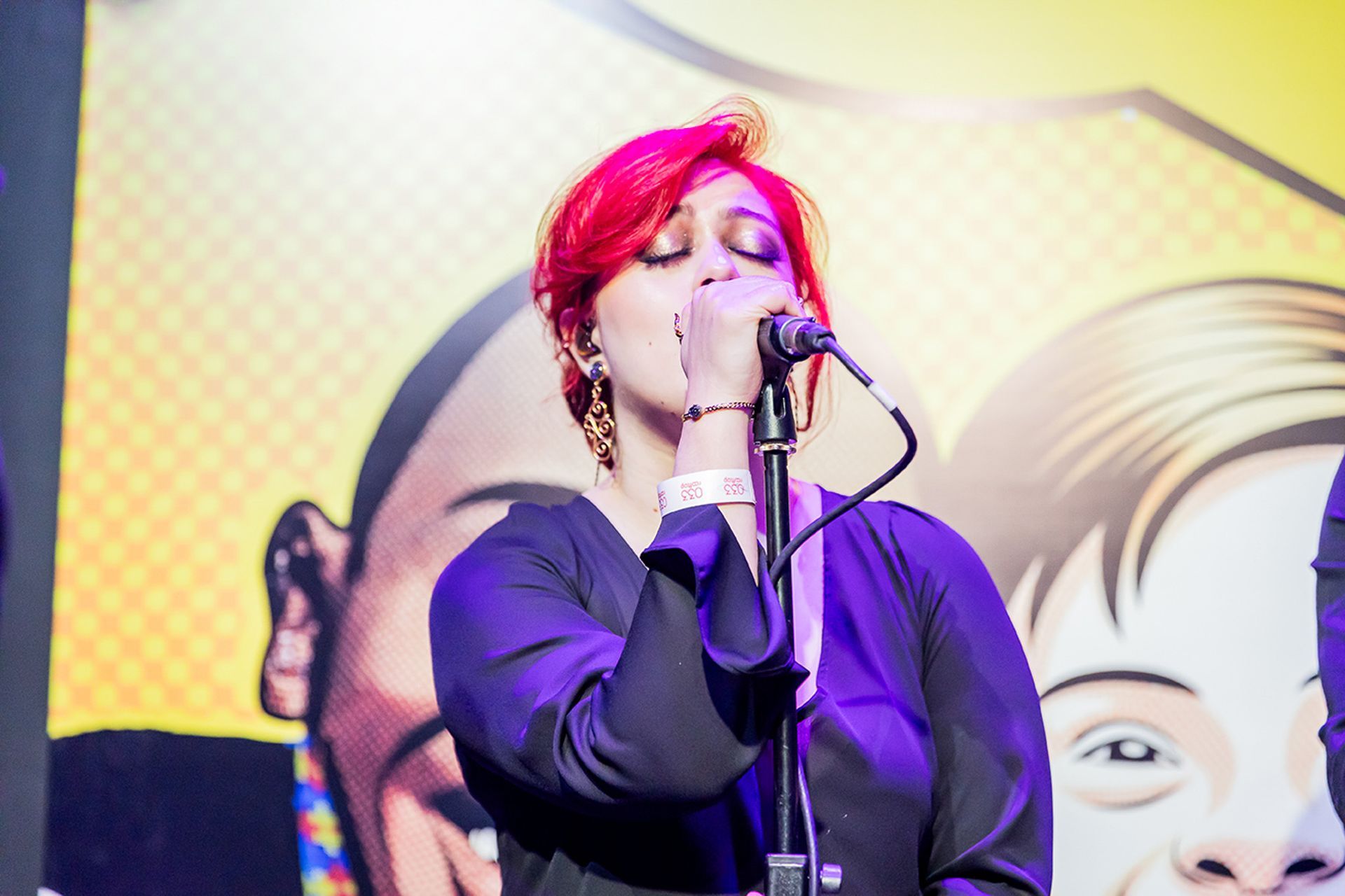 Uma artista com cabelo rosa vibrante canta em um microfone no palco, em frente a um fundo amarelo com ilustrações.