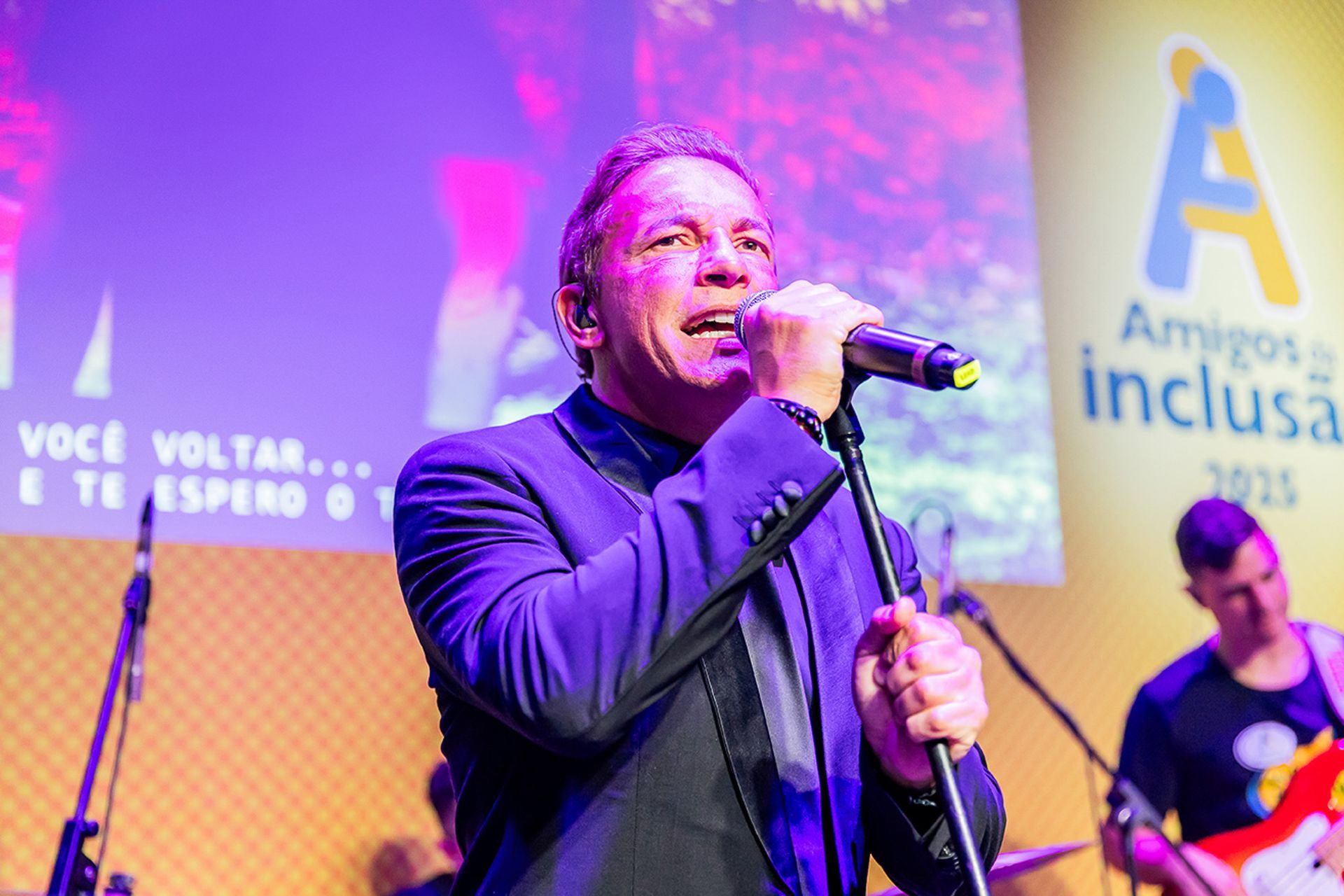 Um cantor de terno escuro se apresenta ao microfone no palco, com o logotipo do evento ao fundo.