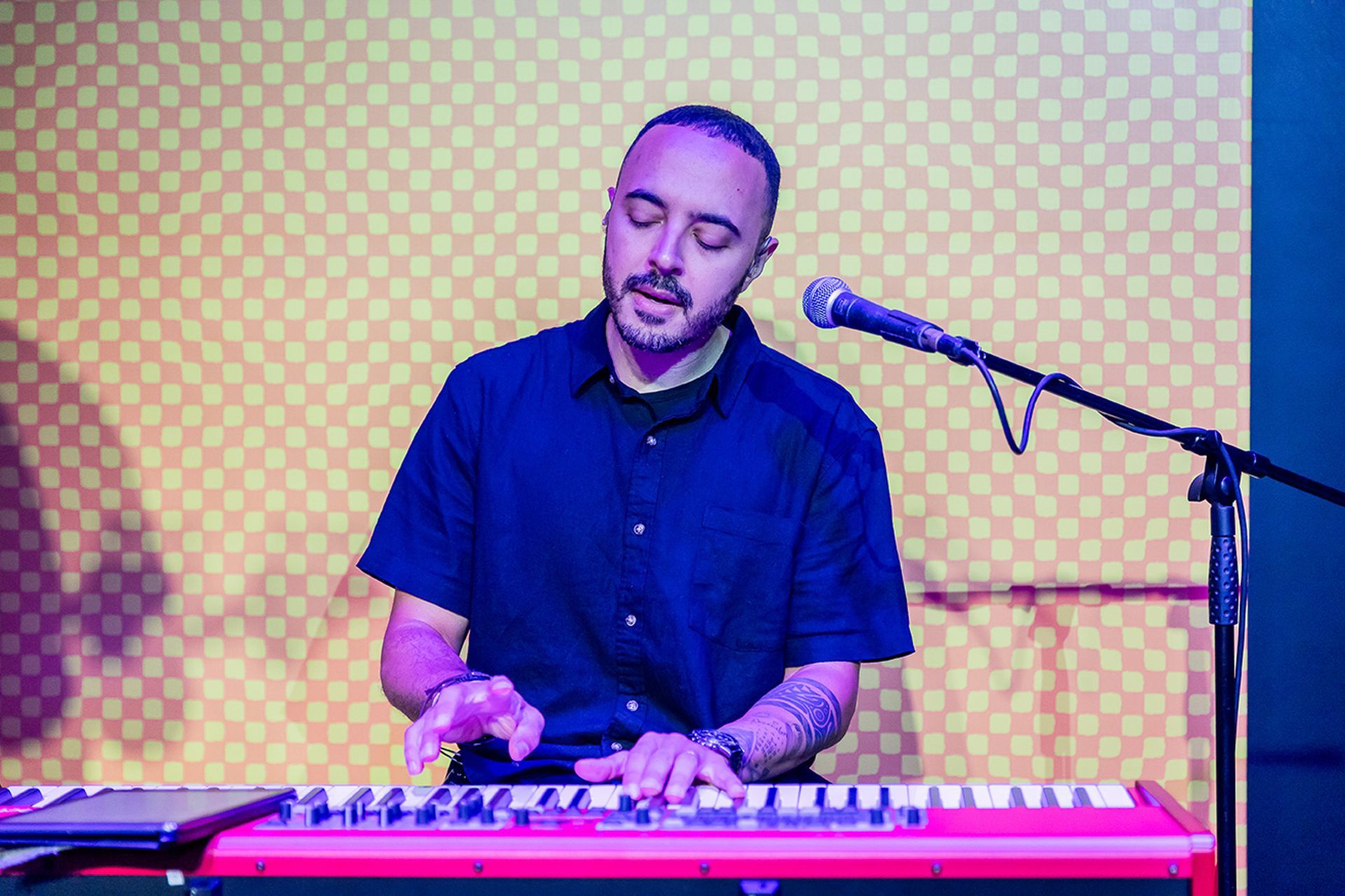 Um artista com barba e tatuagem toca um teclado vermelho em frente a uma parede amarela quadriculada.