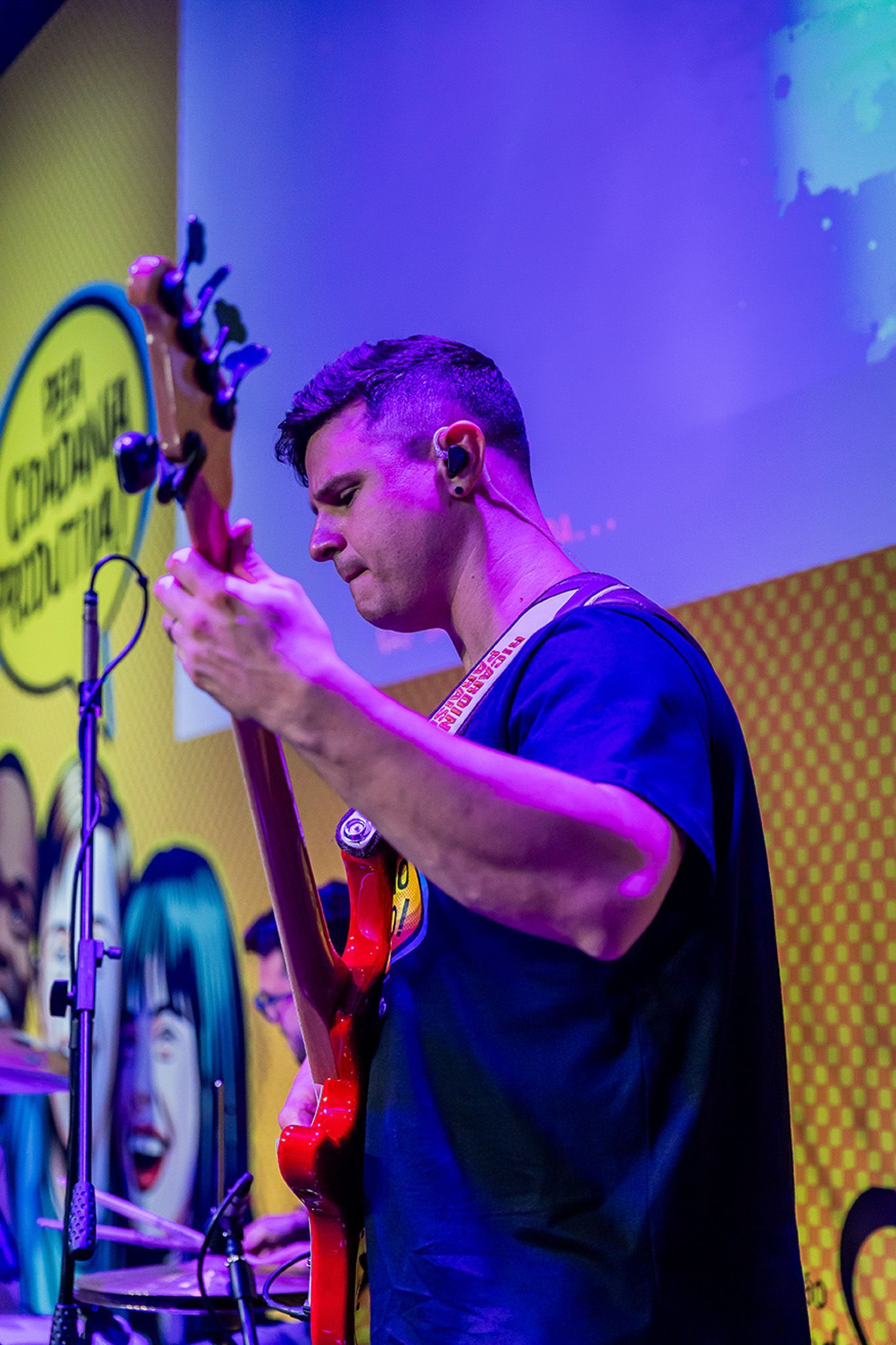 Um músico toca um baixo elétrico vermelho em um palco em frente a um fundo amarelo com rostos ilustrados.