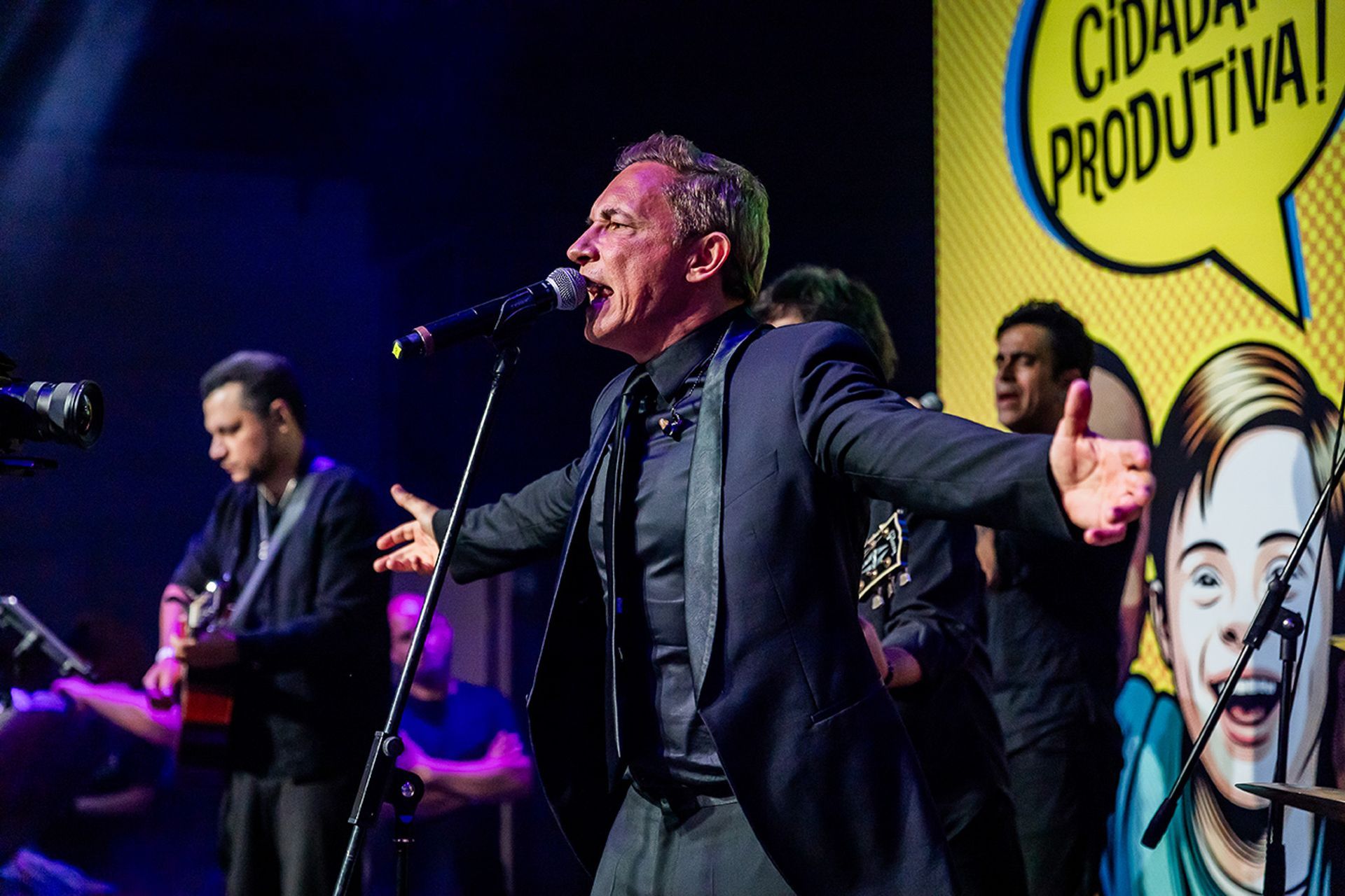 Um artista de terno escuro cantando em um microfone no palco, com uma banda.