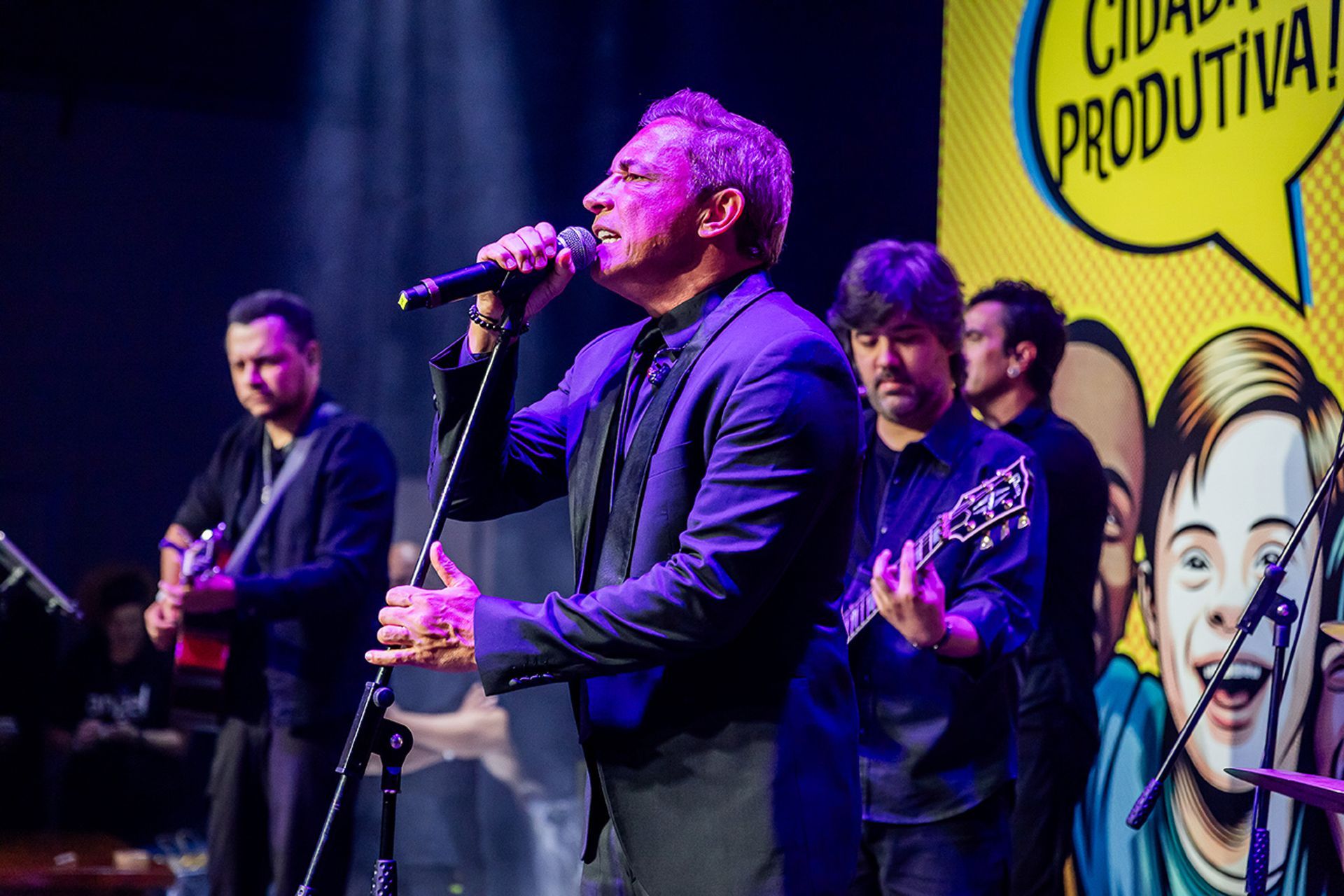 Um artista canta em um microfone no palco, acompanhado por membros da banda que tocam guitarras.