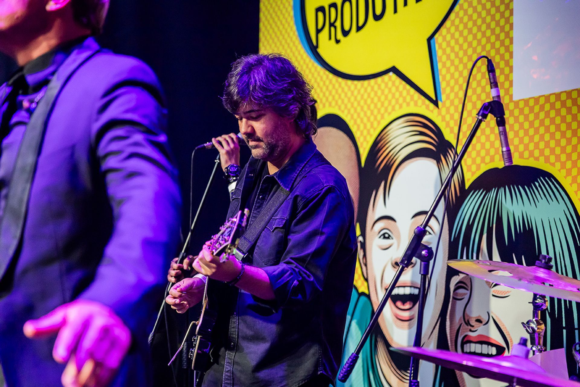Um músico toca guitarra no palco em frente a um cenário colorido, no estilo de desenho animado.