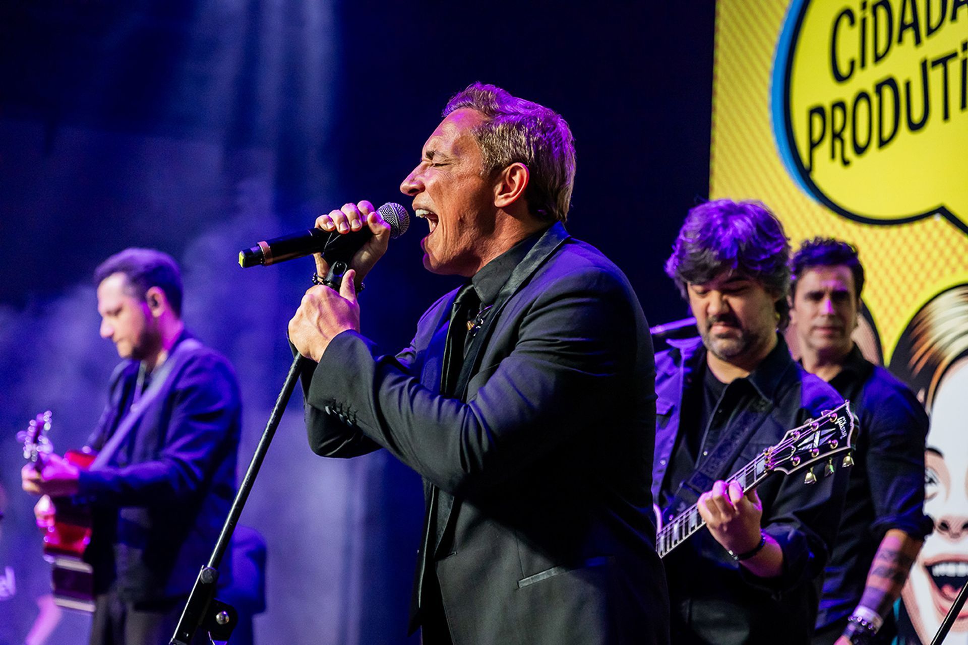 Um artista canta em um microfone no palco, acompanhado por membros da banda que tocam guitarras.