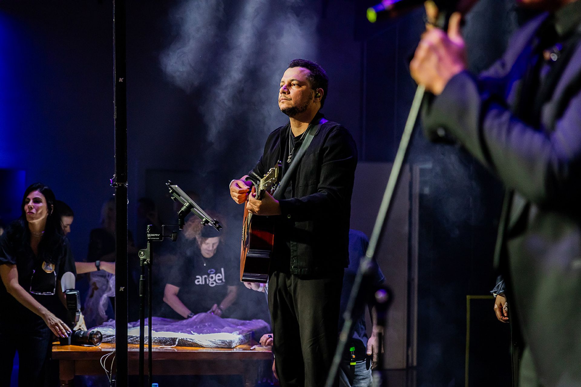 Um músico toca violão no palco, enquanto uma pessoa canta ao microfone em primeiro plano, desfocado.