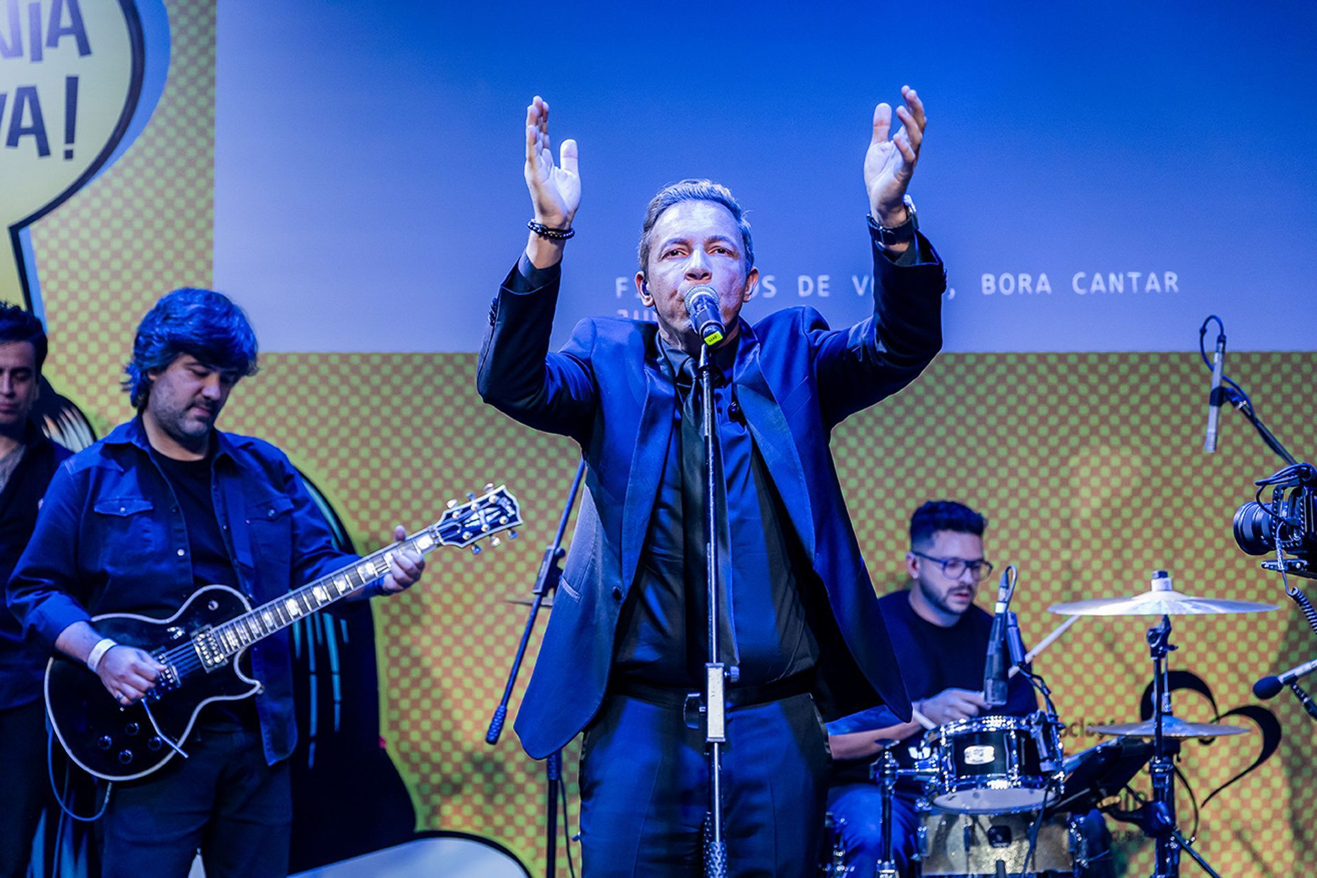 Um cantor no palco, com os braços erguidos, se apresenta com um guitarrista e um baterista contra um fundo amarelo.