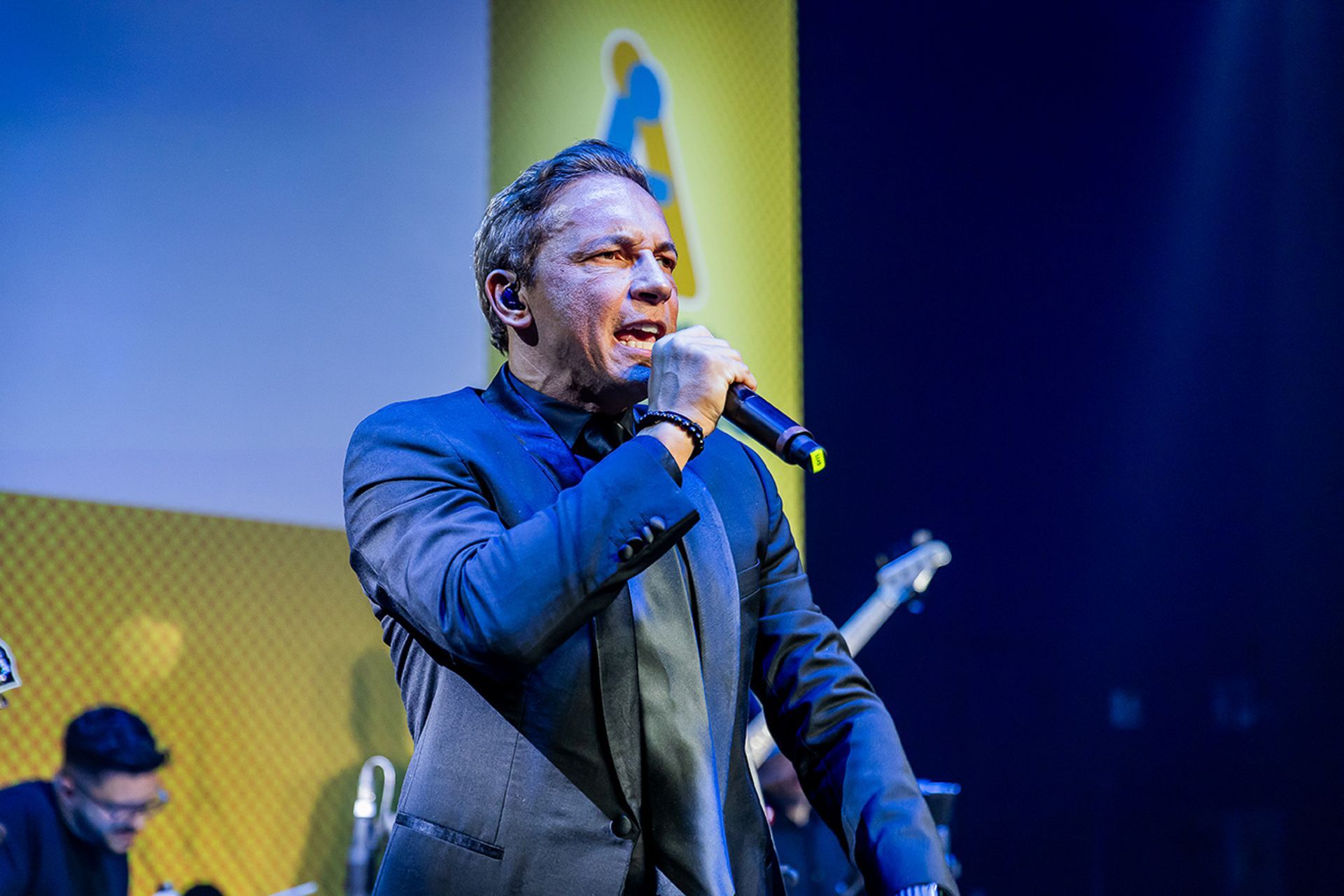 Um artista de paletó canta em um microfone em um palco com fundo amarelo.