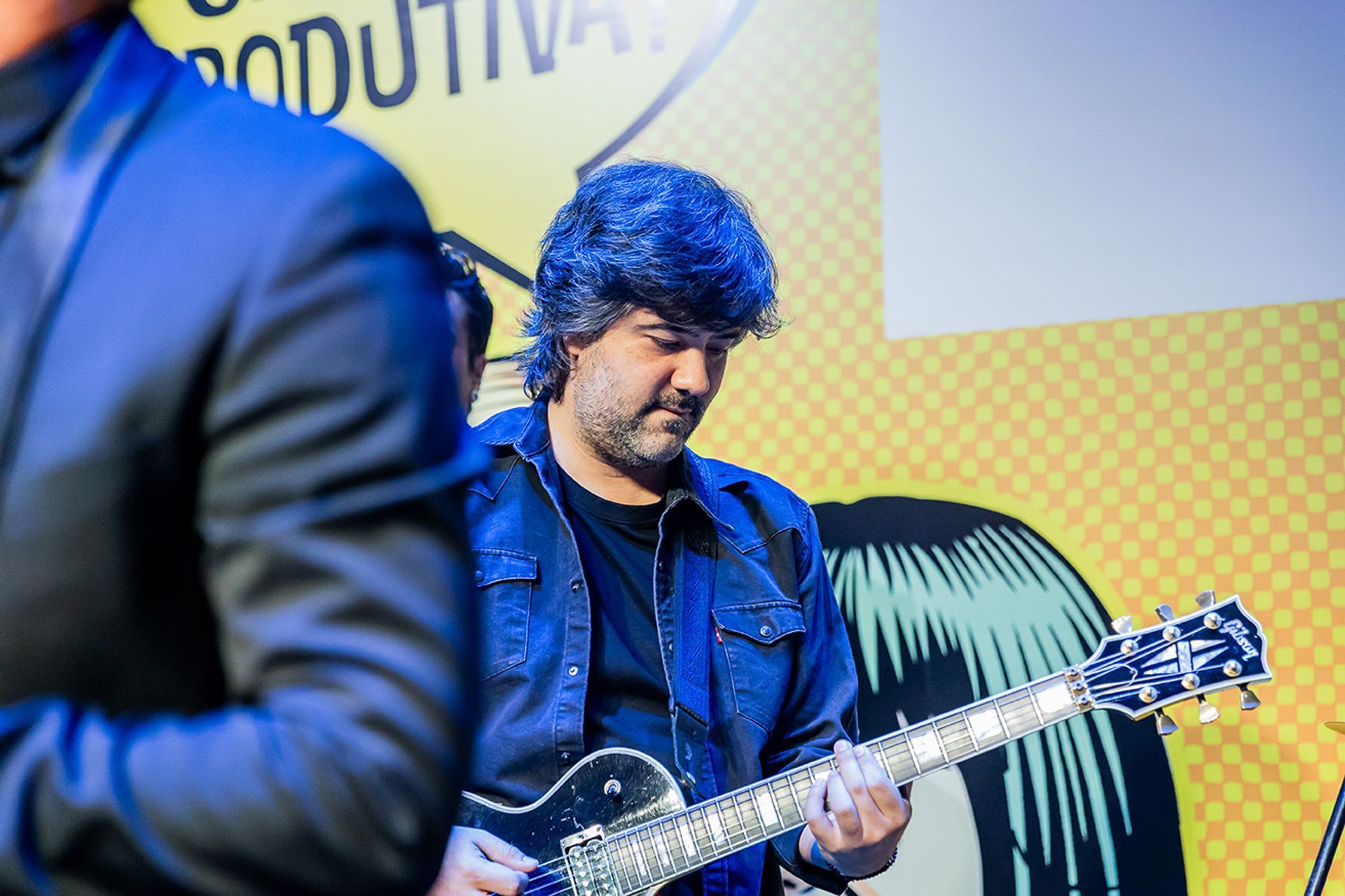 Um artista de cabelos escuros tocando guitarra elétrica contra um fundo amarelo com estilo gráfico de quadrinhos.