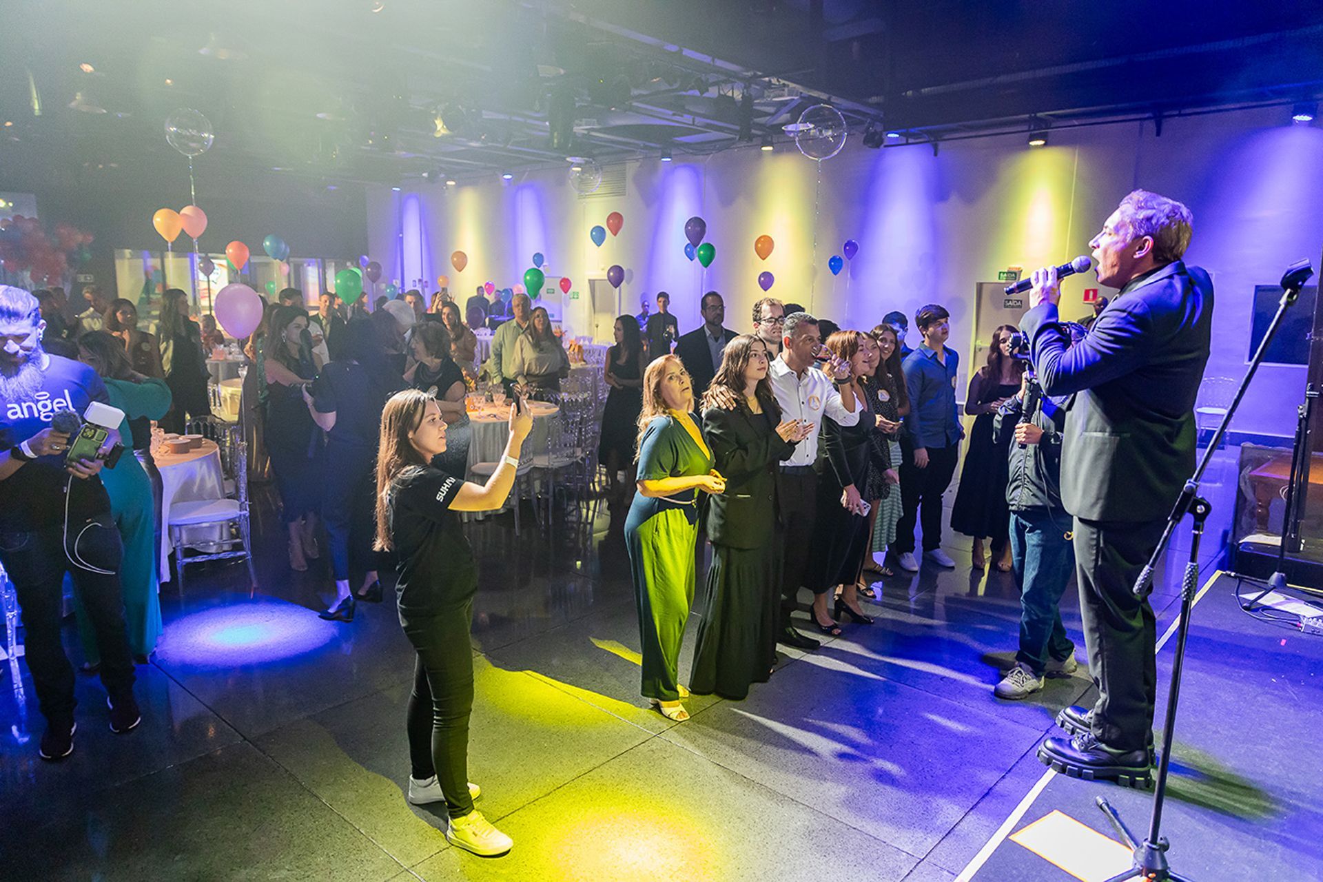 Um artista canta ao microfone para uma plateia em um salão de eventos com iluminação tênue, decorado com balões e luzes.