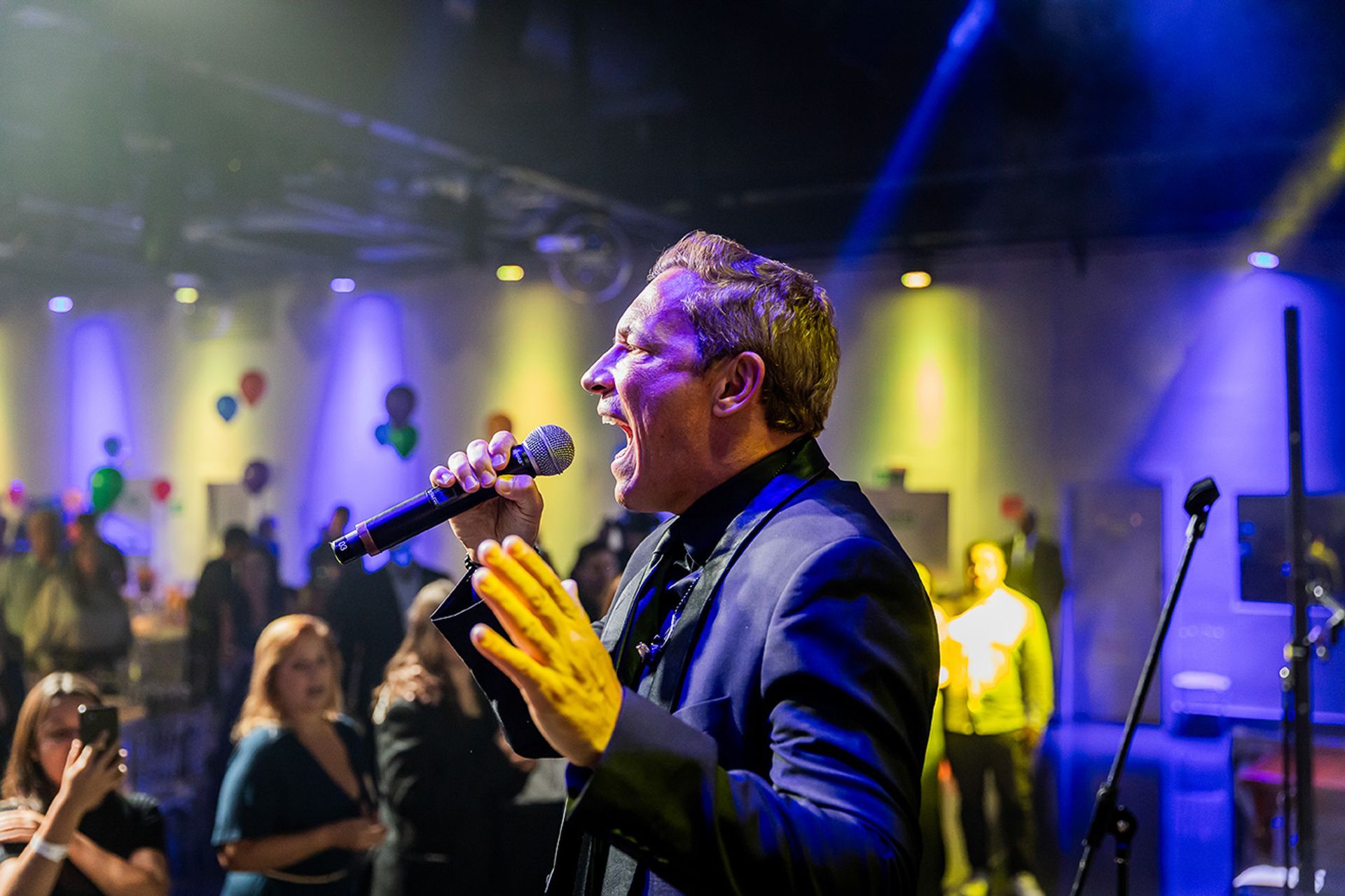 Um cantor se apresenta ao microfone no palco, iluminada por holofotes azuis e amarelos em um espaço para eventos.