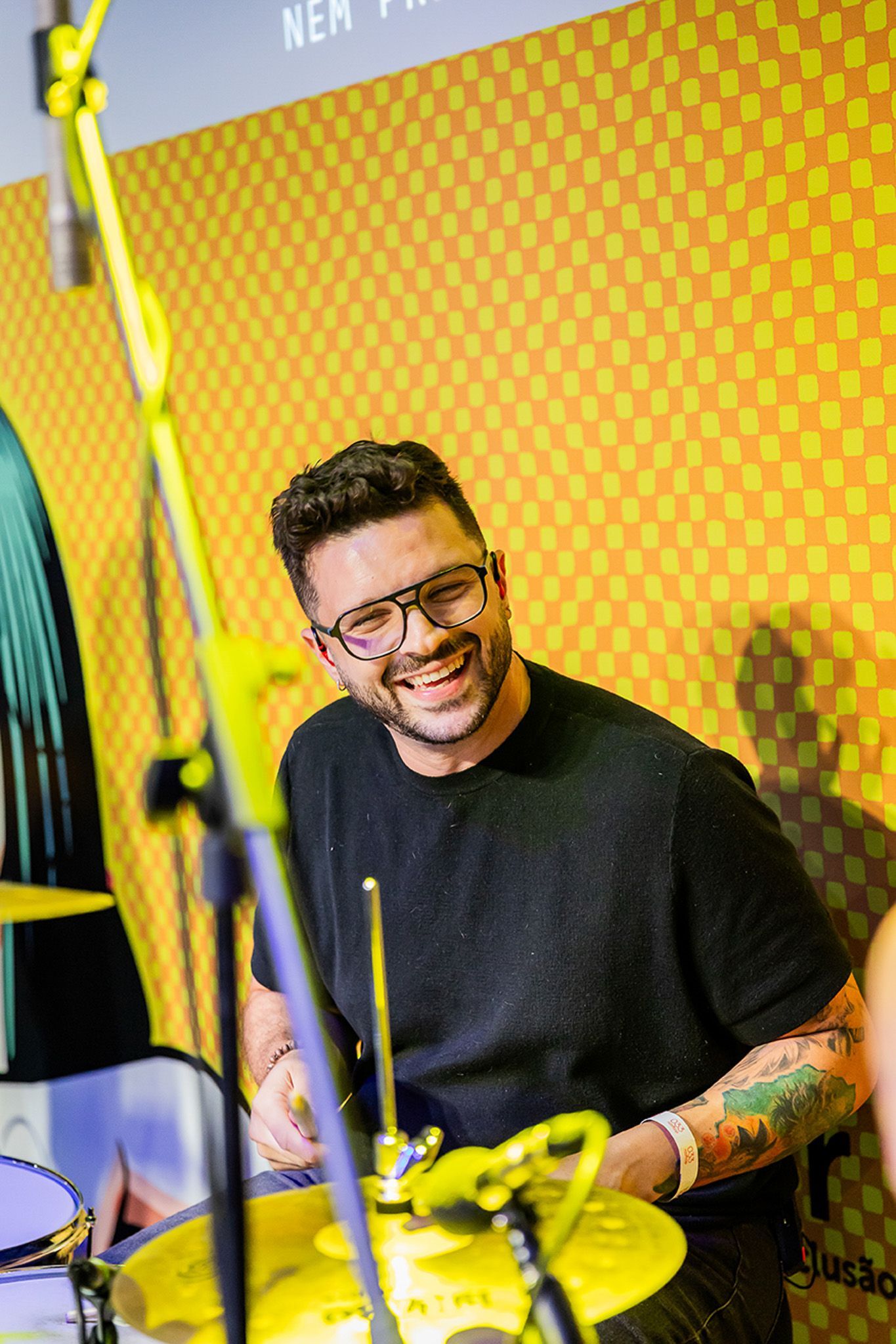 Um artista sorridente, de óculos e com tatuagens no braço, toca bateria em frente a uma parede amarela com estampa.