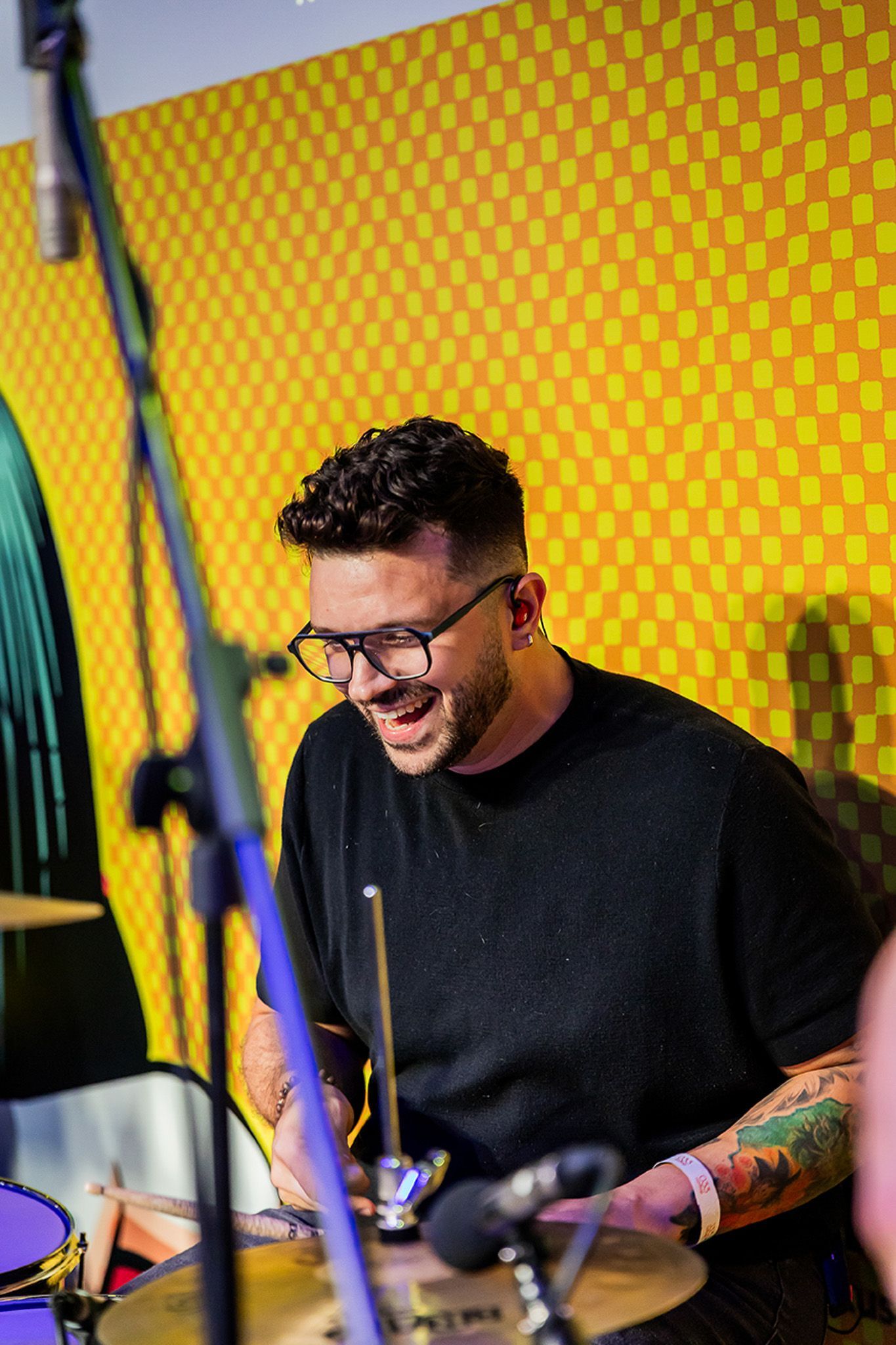 Um músico sorridente com tatuagens no braço toca bateria em frente a uma parede quadriculada amarela e laranja.