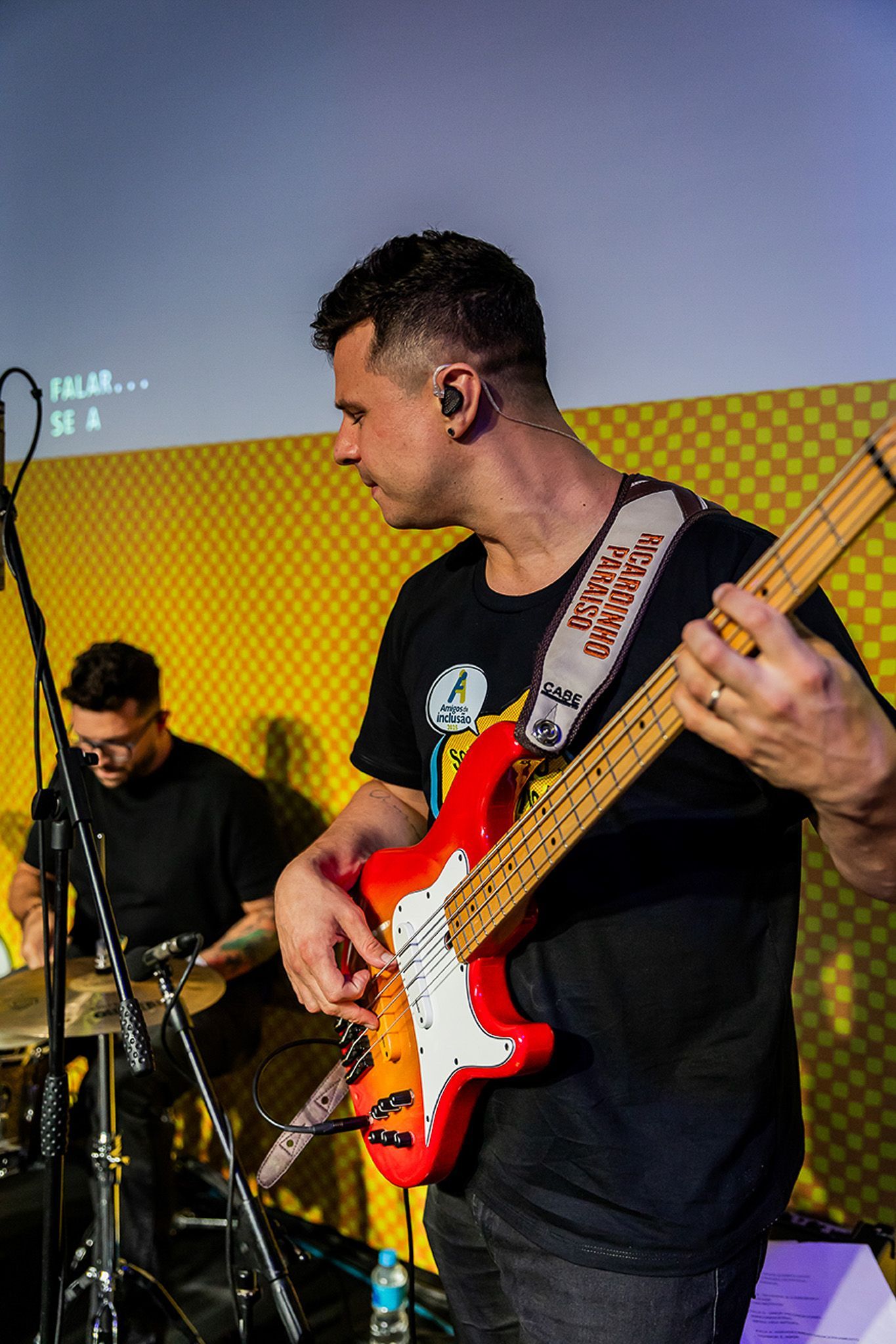 Um músico tocando um baixo elétrico vermelho brilhante em um estúdio com uma parede amarela quadriculada.