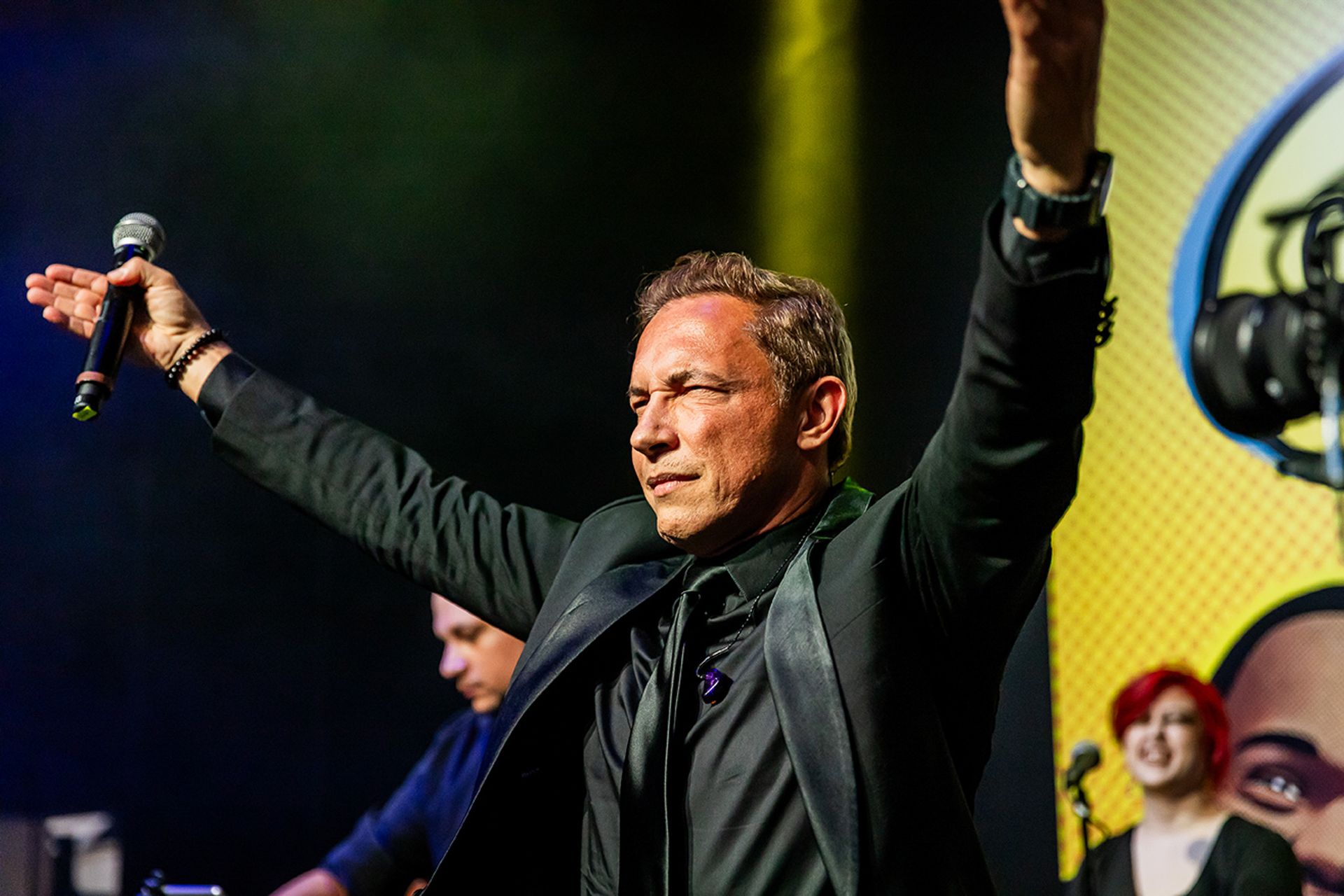 Um artista de terno preto no palco, com os braços erguidos, segurando um microfone, com músicos ao fundo.