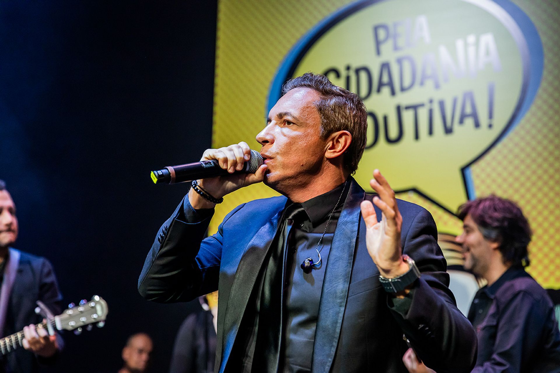 Um artista de terno preto canta em um microfone no palco, com uma faixa atrás que diz 