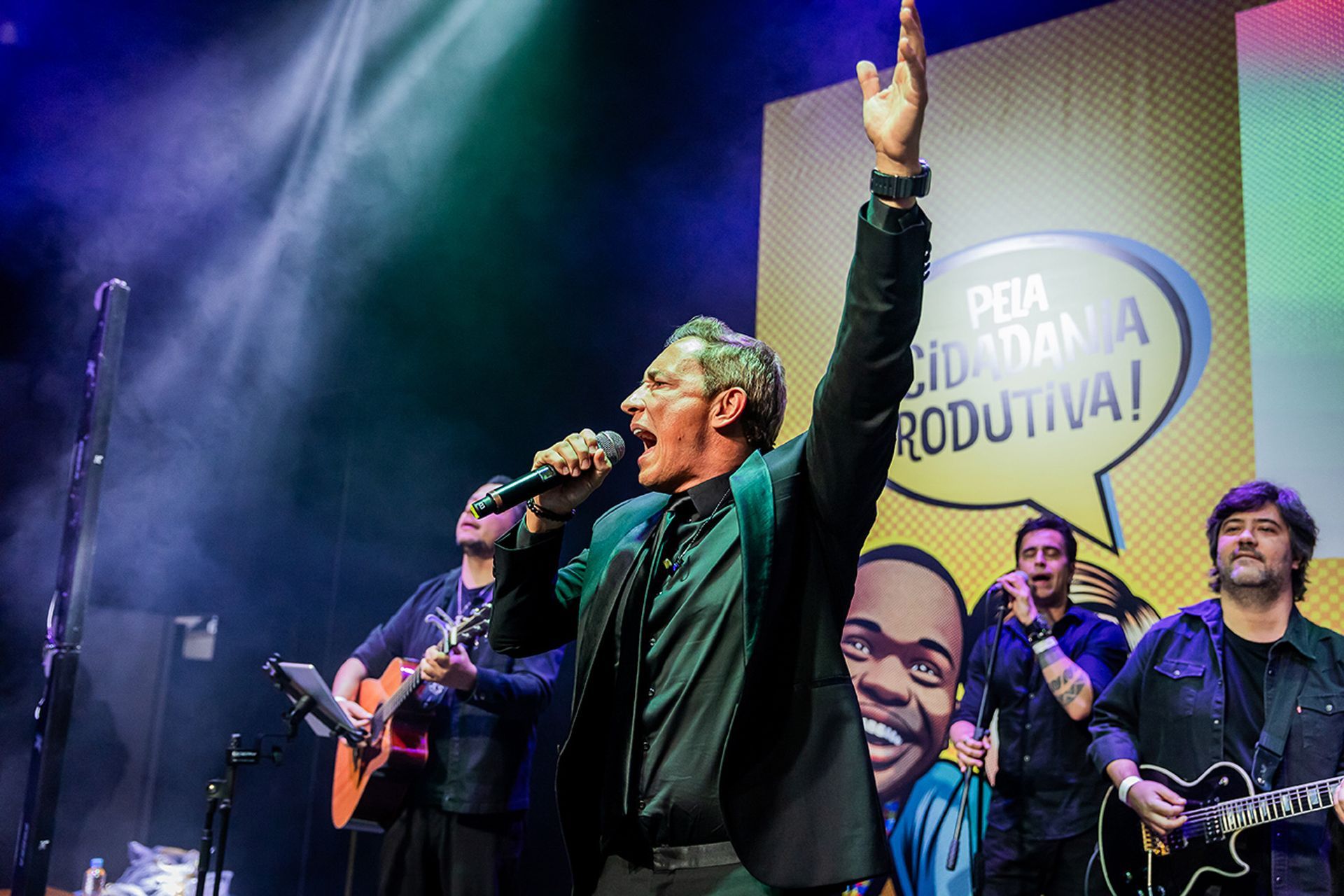 Um artista de blazer verde canta ao microfone com o braço erguido no palco, acompanhado por músicos e um cartaz de campanha.
