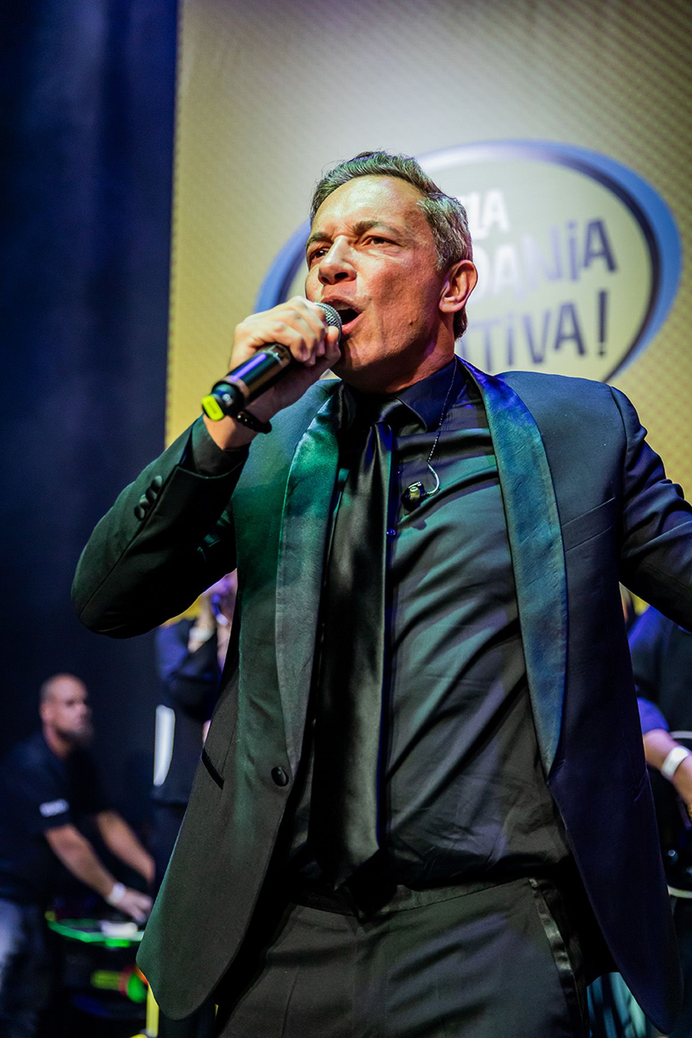 Um artista de terno escuro com lapelas iridescentes canta em um microfone em um palco com uma placa de fundo amarela.