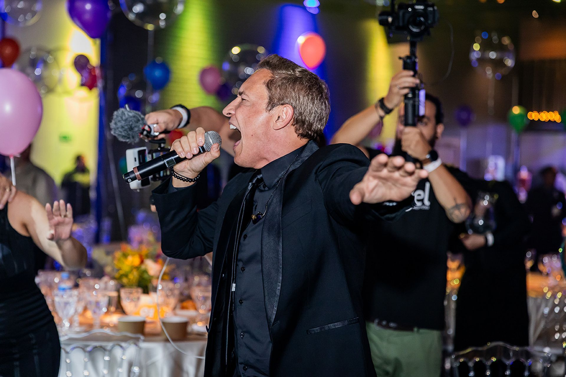 Um artista de terno preto canta com paixão em um microfone durante um evento, enquanto um cinegrafista filma ao fundo.