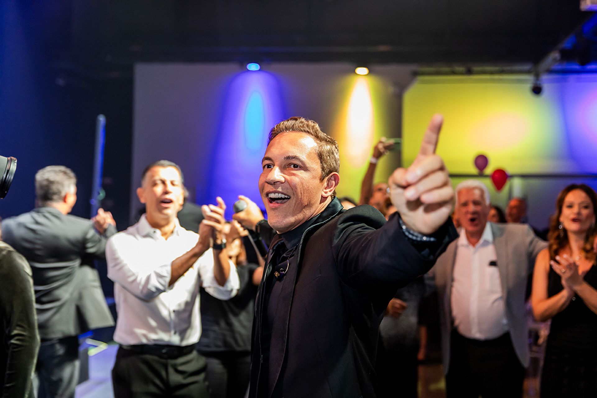 Um artista sorridente, vestindo um paletó, gesticula para cima enquanto comemora com um grupo diversificado de pessoas.