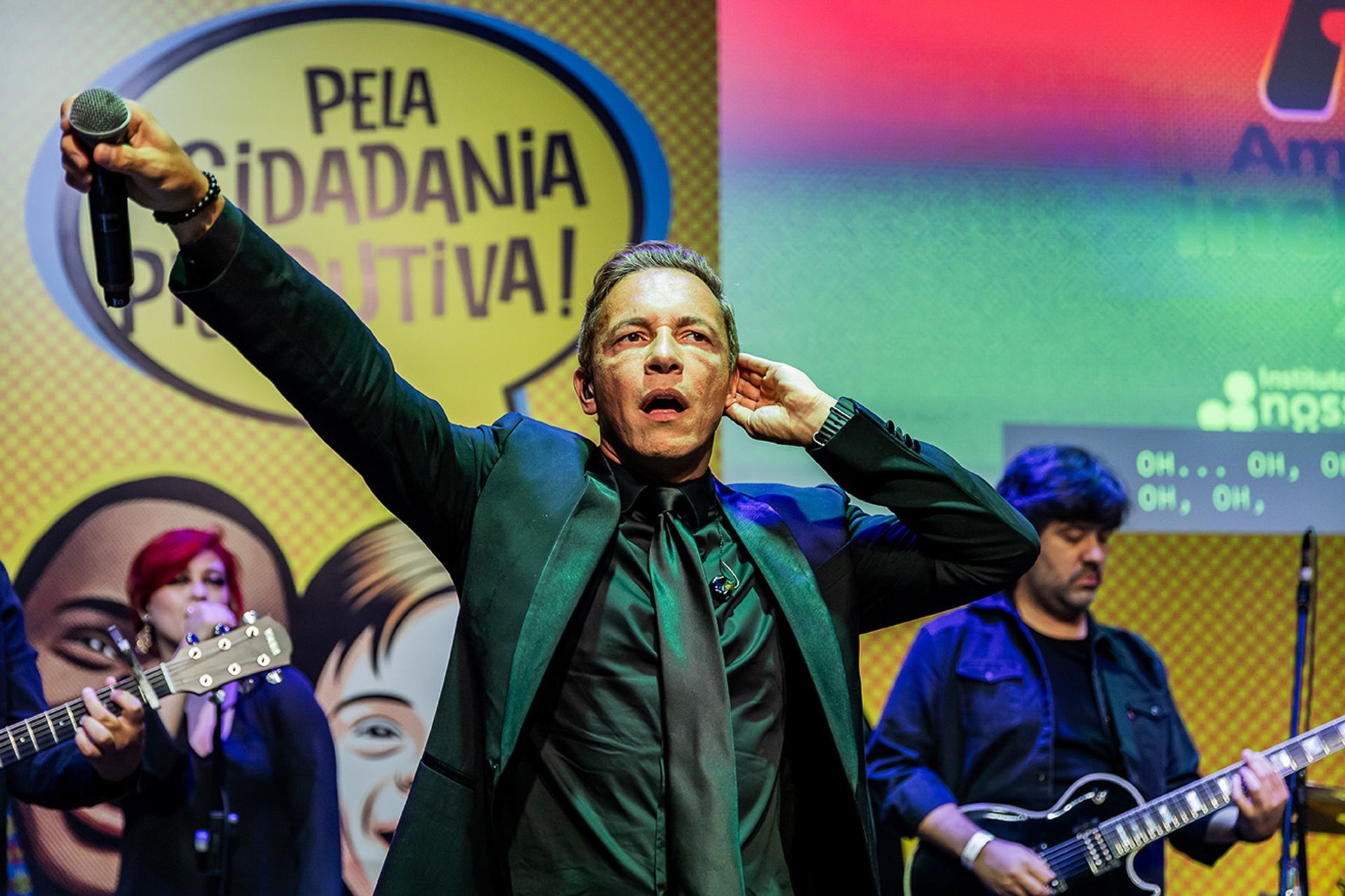 Um artista de blazer verde canta em um microfone no palco, com um músico tocando violão.