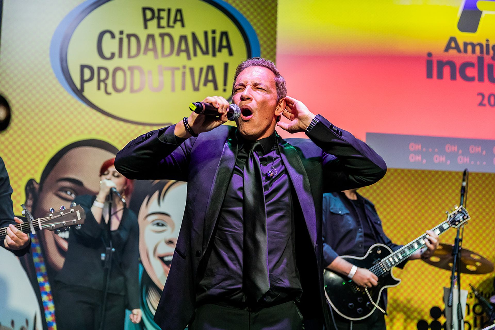Um artista de terno escuro canta em um microfone no palco, com os membros da banda e um cenário vibrante do evento.