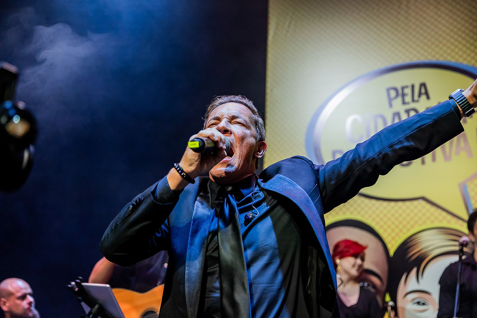 Um artista de terno azul canta em um microfone no palco, gesticulando para uma plateia.