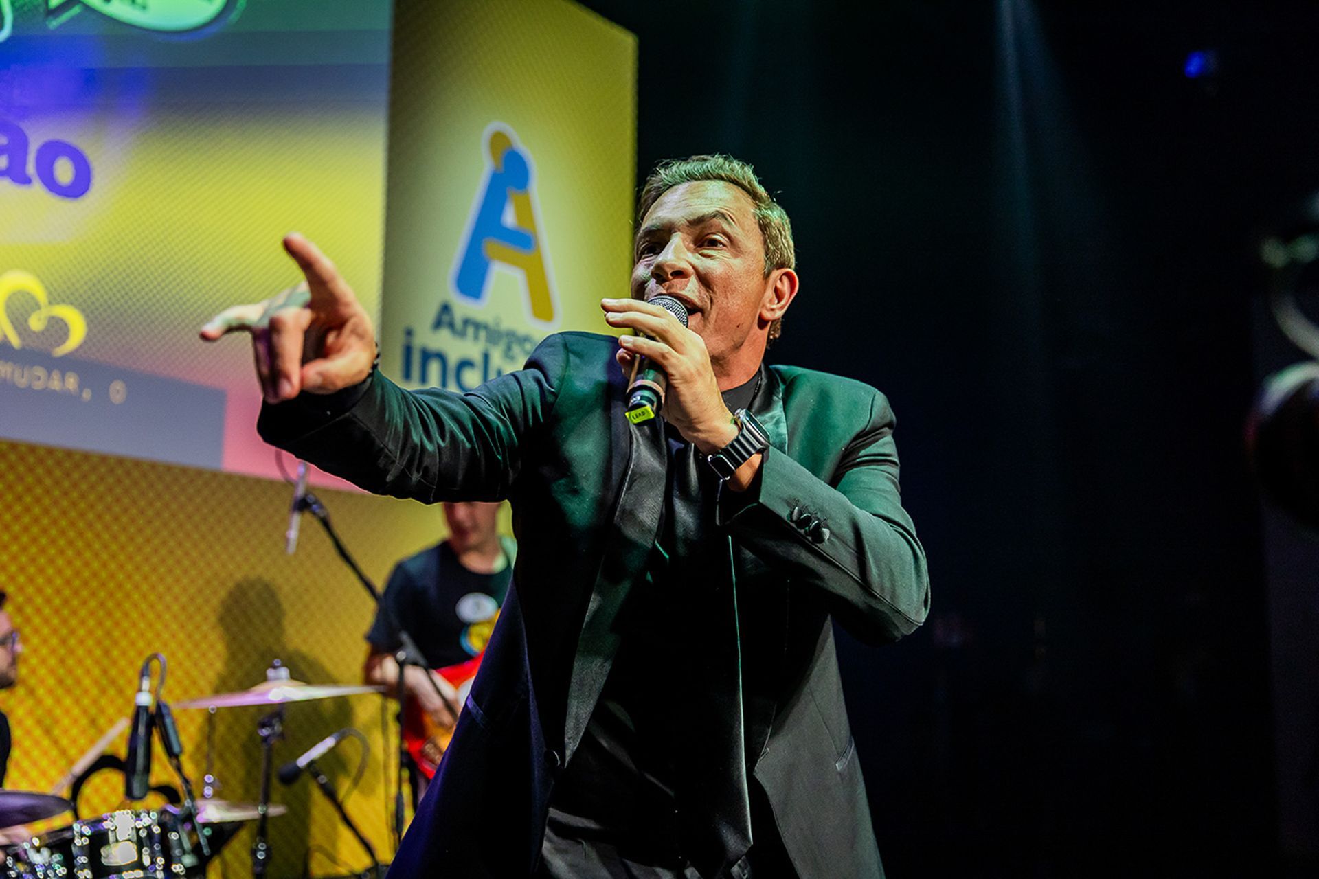 Um artista de terno escuro aponta enquanto canta em um microfone no palco, com uma faixa atrás dele em um local iluminado.
