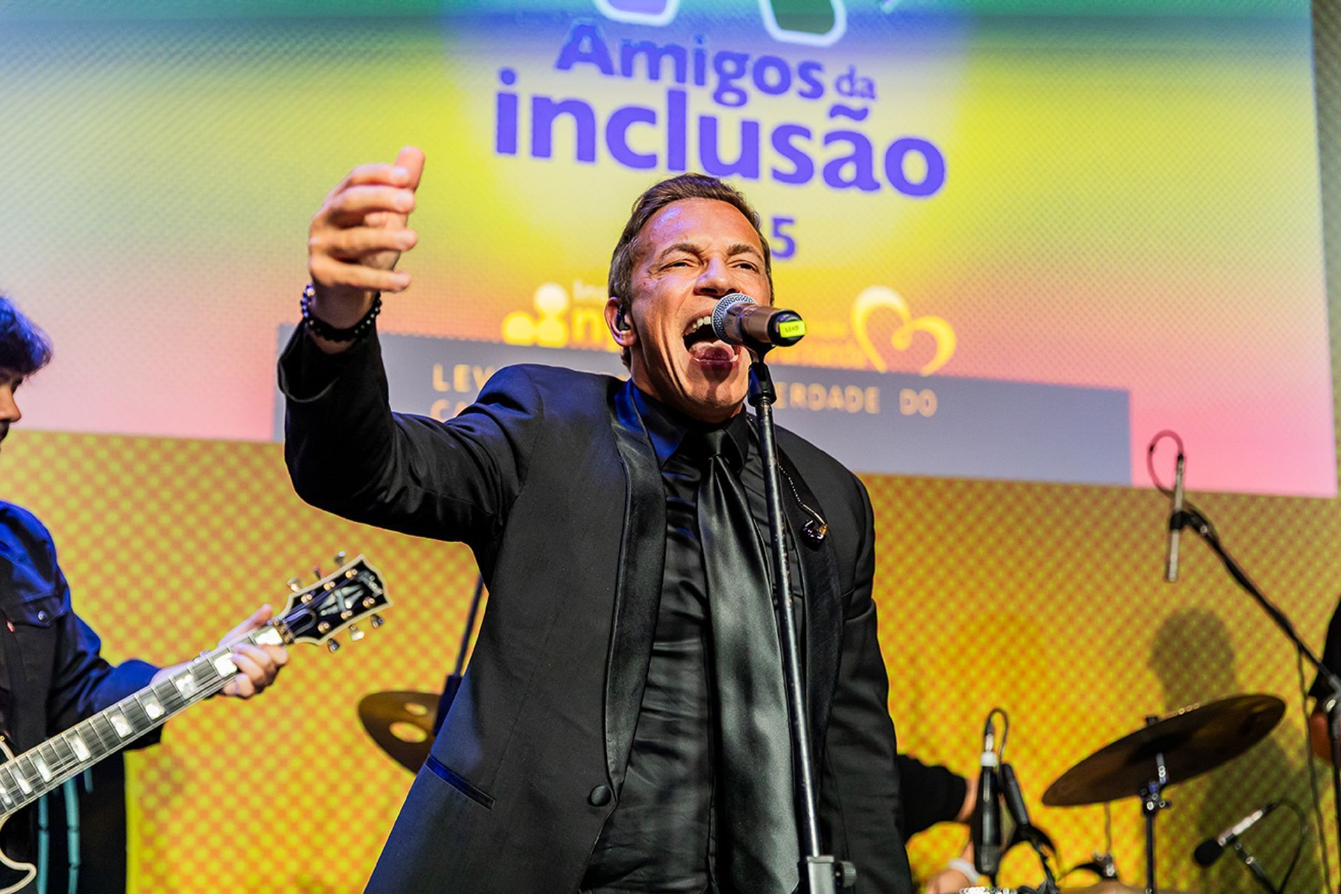 Um cantor de terno se apresenta no palco em frente a uma faixa com os dizeres 