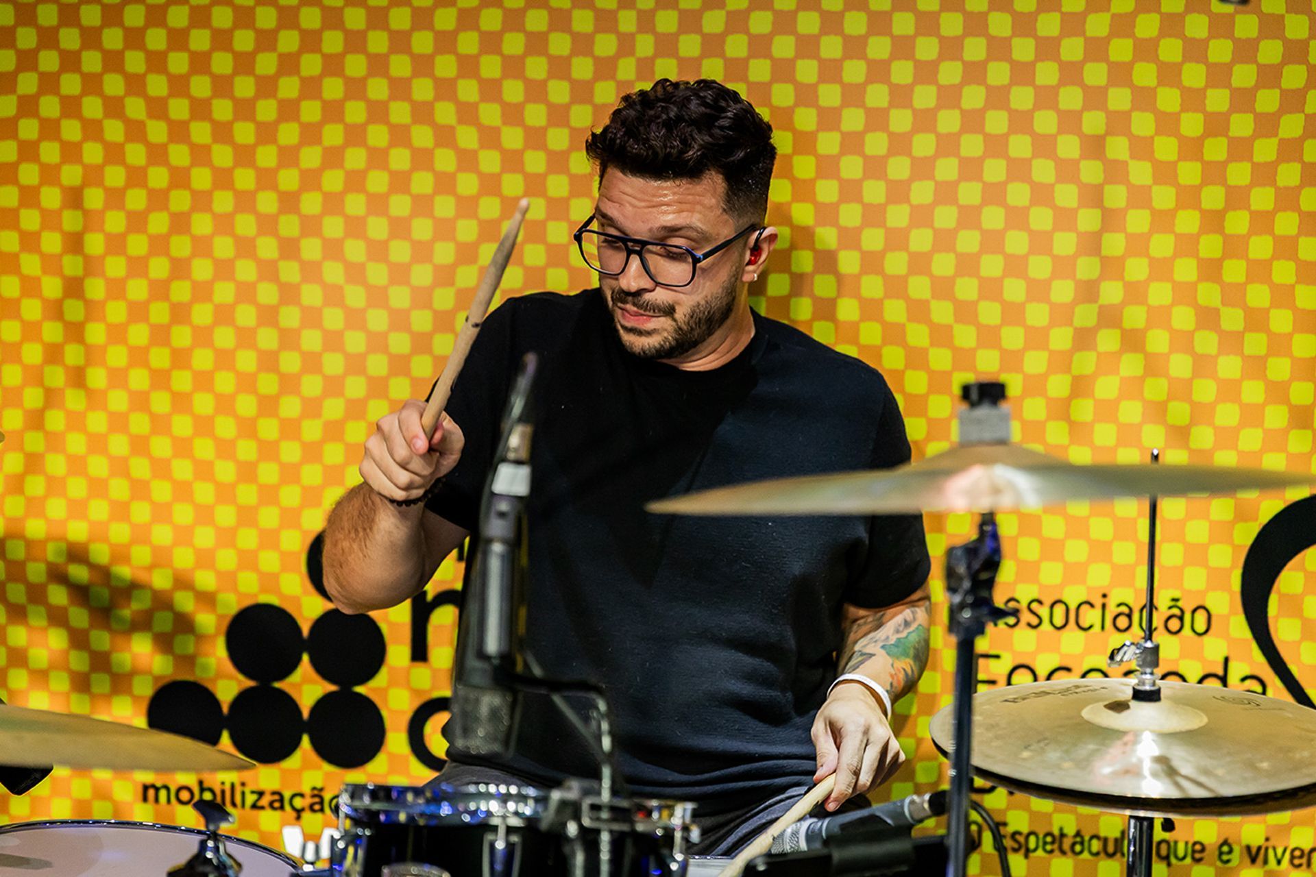 Um artista tocando bateria, usando óculos e uma camiseta preta, em um fundo amarelo com padrões circulares.