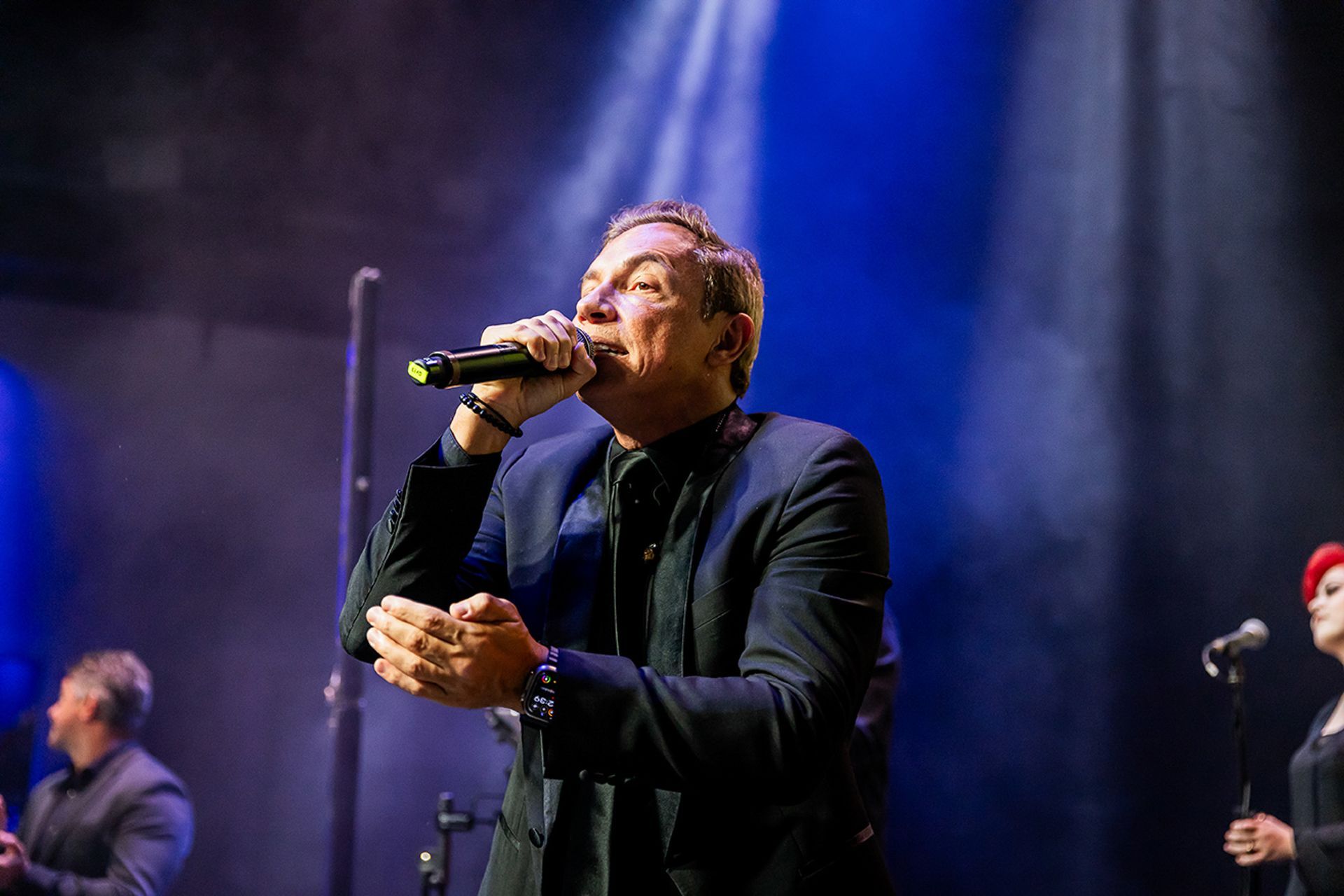 Um artista de terno escuro canta em um microfone no palco, iluminado por feixes de luz azul brilhante.