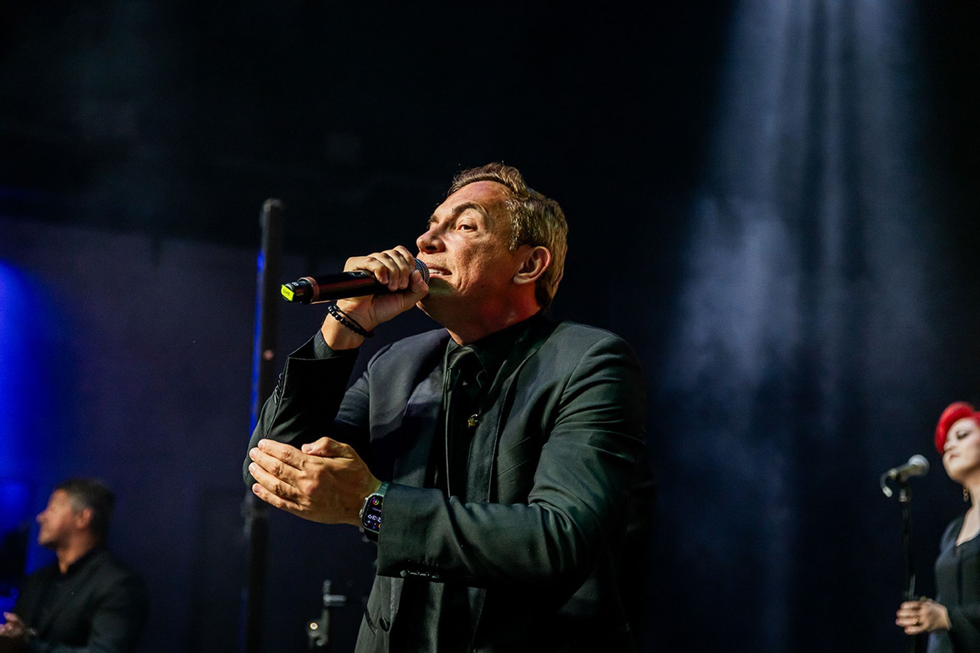 Um artista de terno preto canta em um microfone em um palco com iluminação dramática e uma vocalista de apoio próxima.