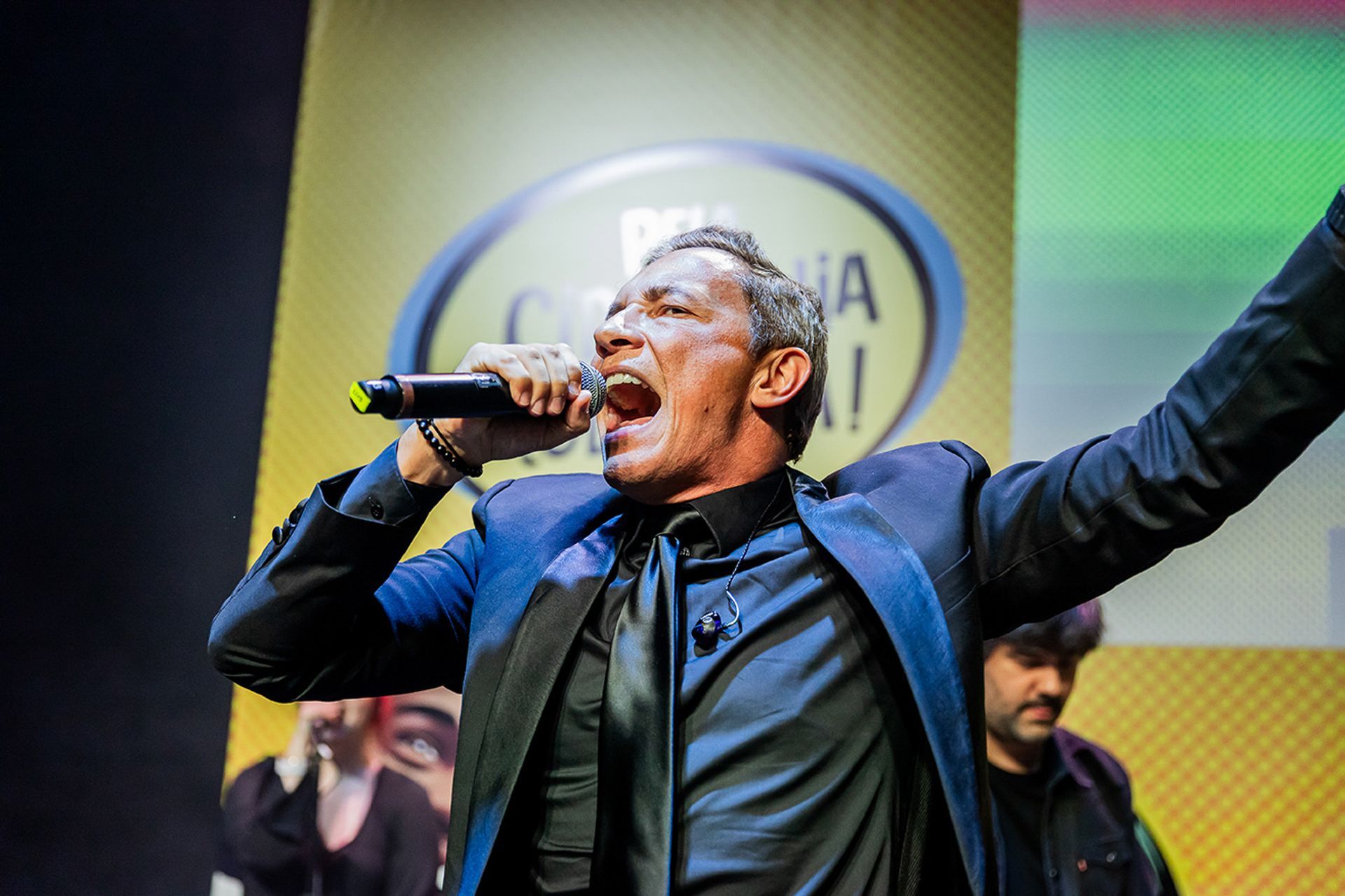 Um artista de terno escuro cantando em um microfone no palco, com um logotipo oval ao fundo.