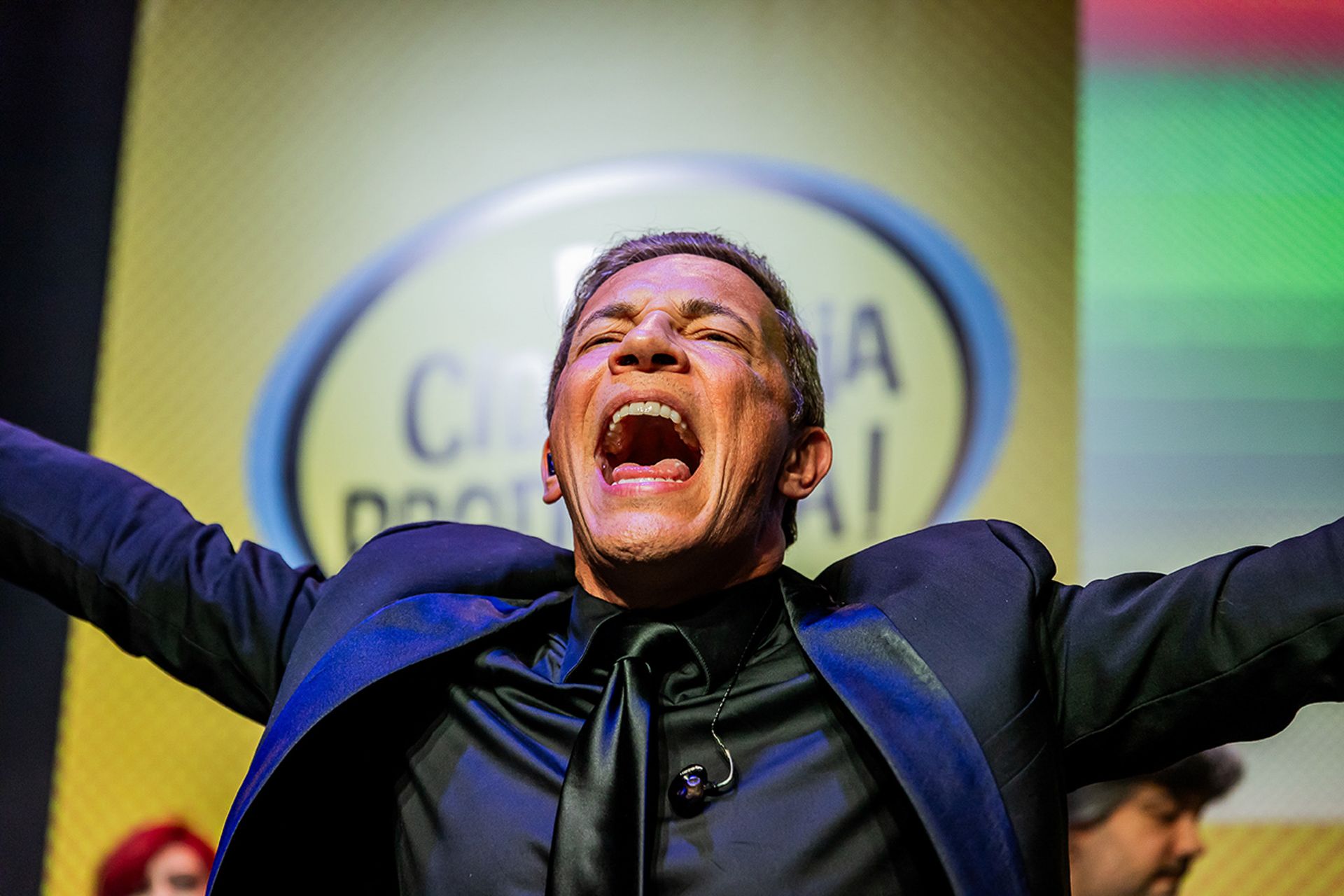 Um artista de terno e gravata está de pé, com os braços estendidos e a boca aberta, em uma pose expressiva e enérgica no palco.