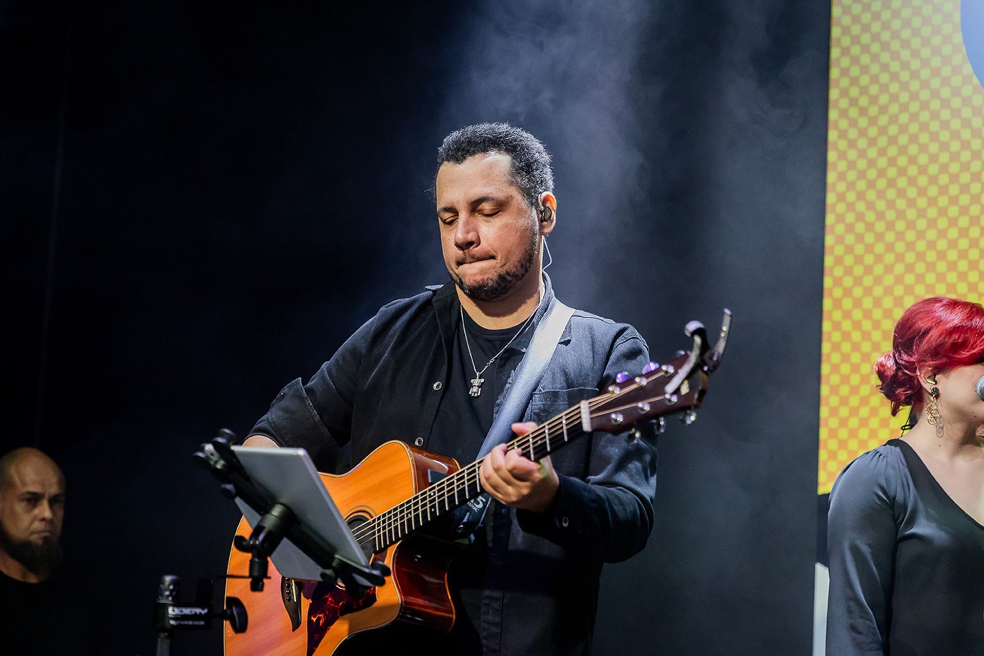 Um músico toca violão no palco, com o foco em uma estante de partituras, enquanto outros músicos são visíveis nas laterais.