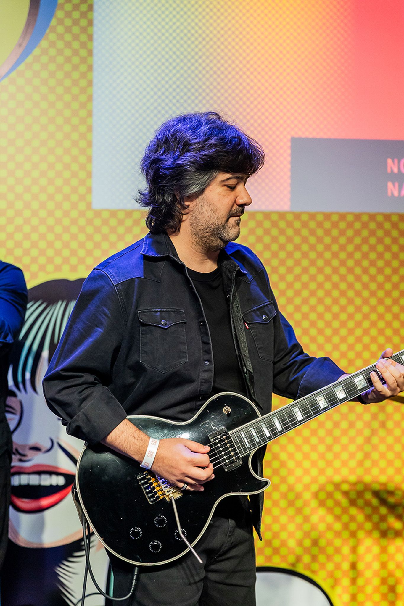 Um músico de cabelos escuros e cacheados tocando uma guitarra elétrica preta em frente a um fundo amarelo estampado.