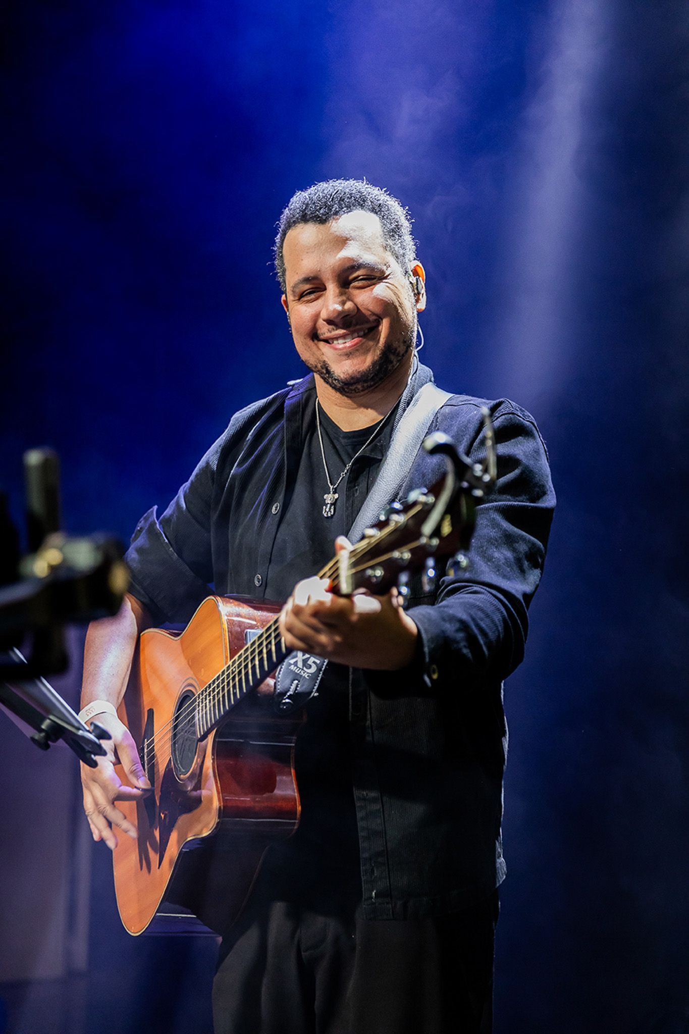 Um músico sorridente tocando violão em um palco sob holofotes azuis.