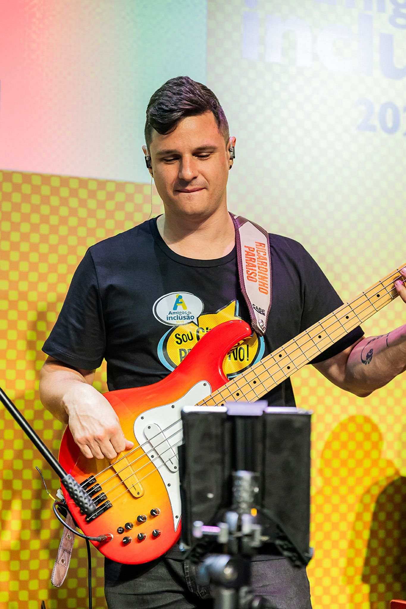 Um músico de cabelo curto toca um baixo elétrico vermelho e amarelo no palco, em frente a um cenário amarelo estampado.