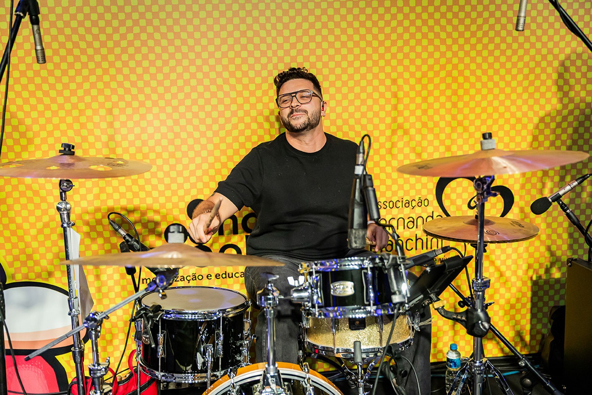 Um baterista tocando bateria em um estúdio, contra uma parede amarela com estampa.