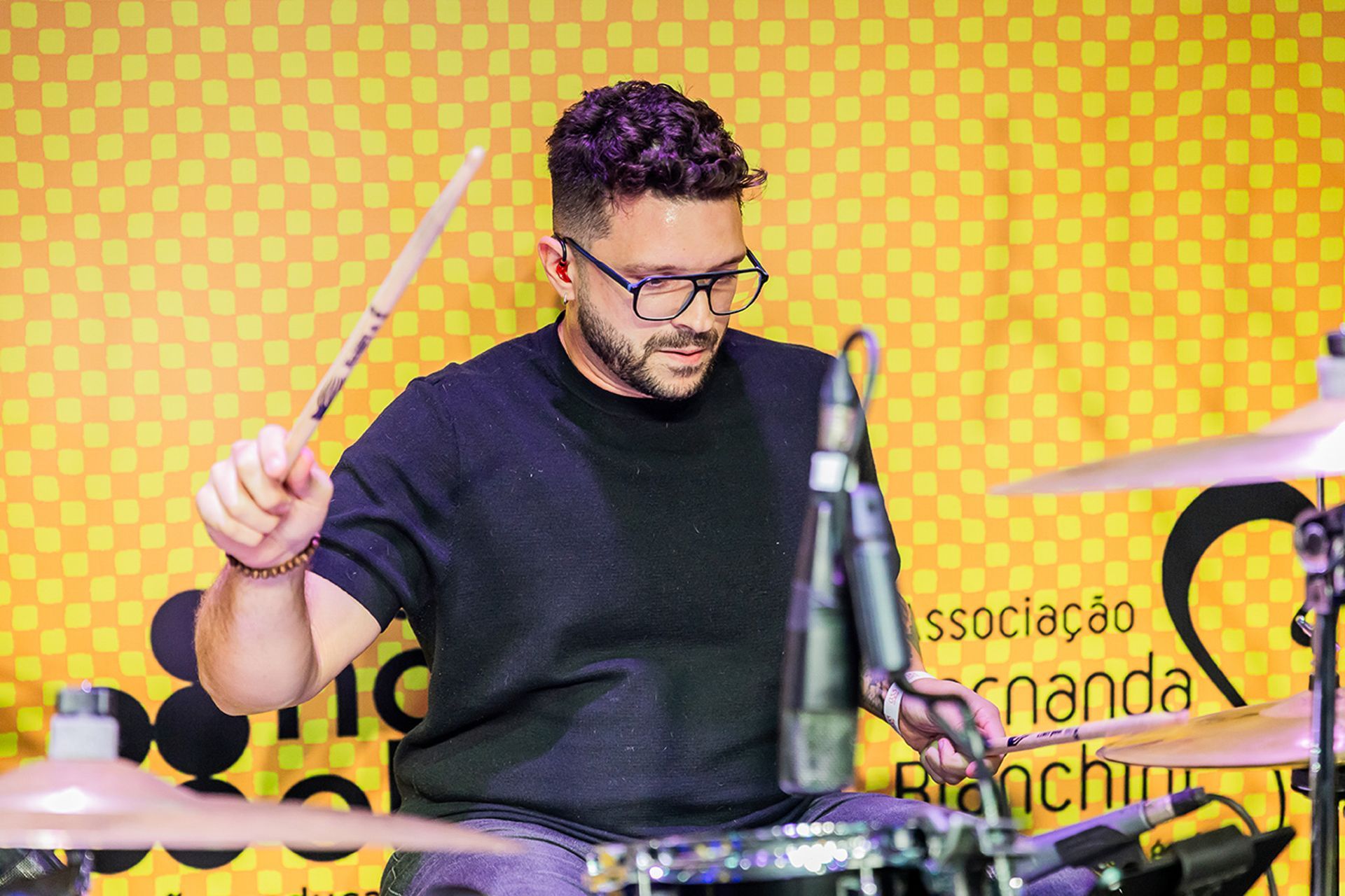 Um artista de óculos e camisa escura toca bateria em frente a um fundo xadrez amarelo com texto.
