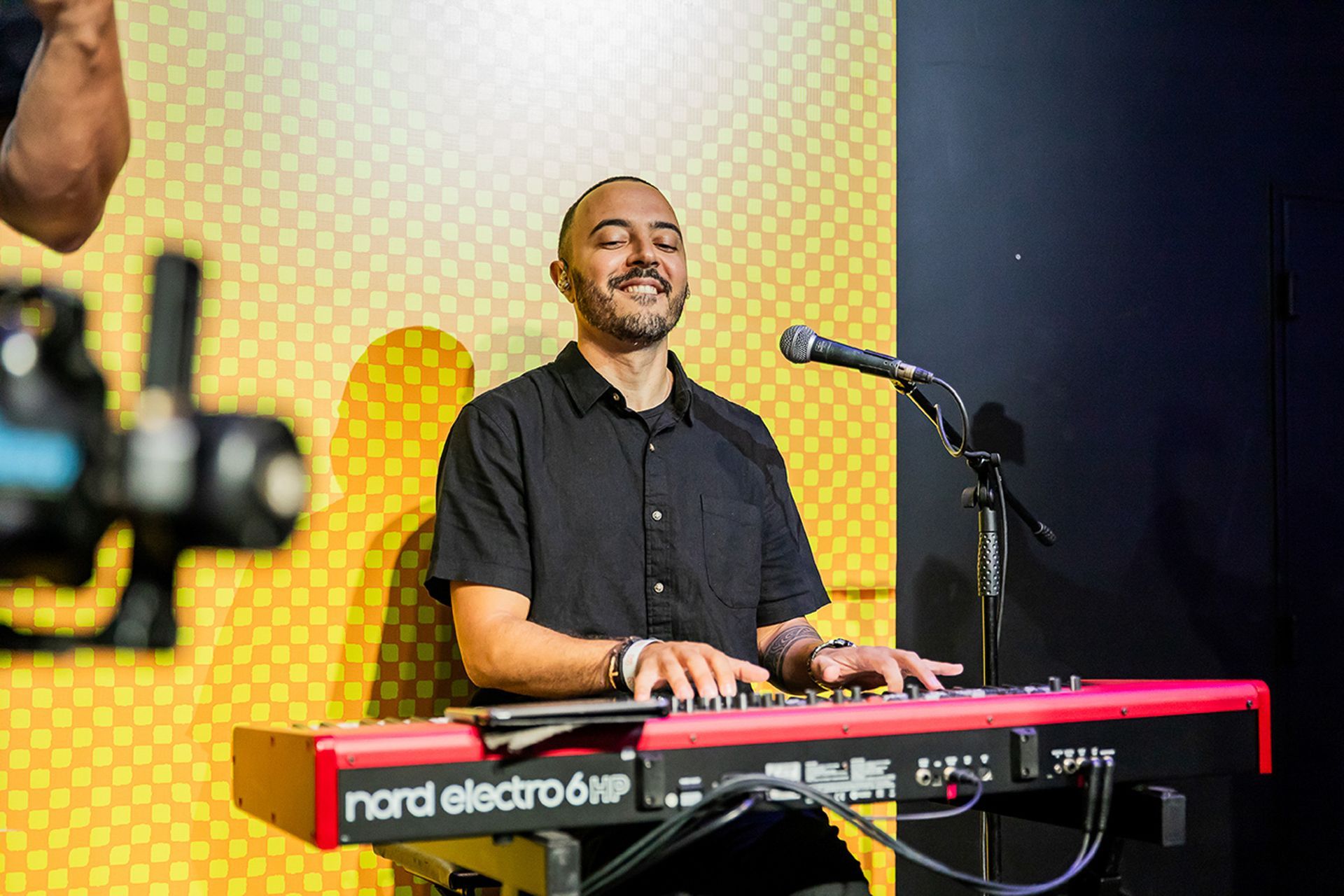 Um artista barbuda sorri enquanto toca num teclado HP Nord Electro 6 vermelho em frente a uma parede amarela com padrões.