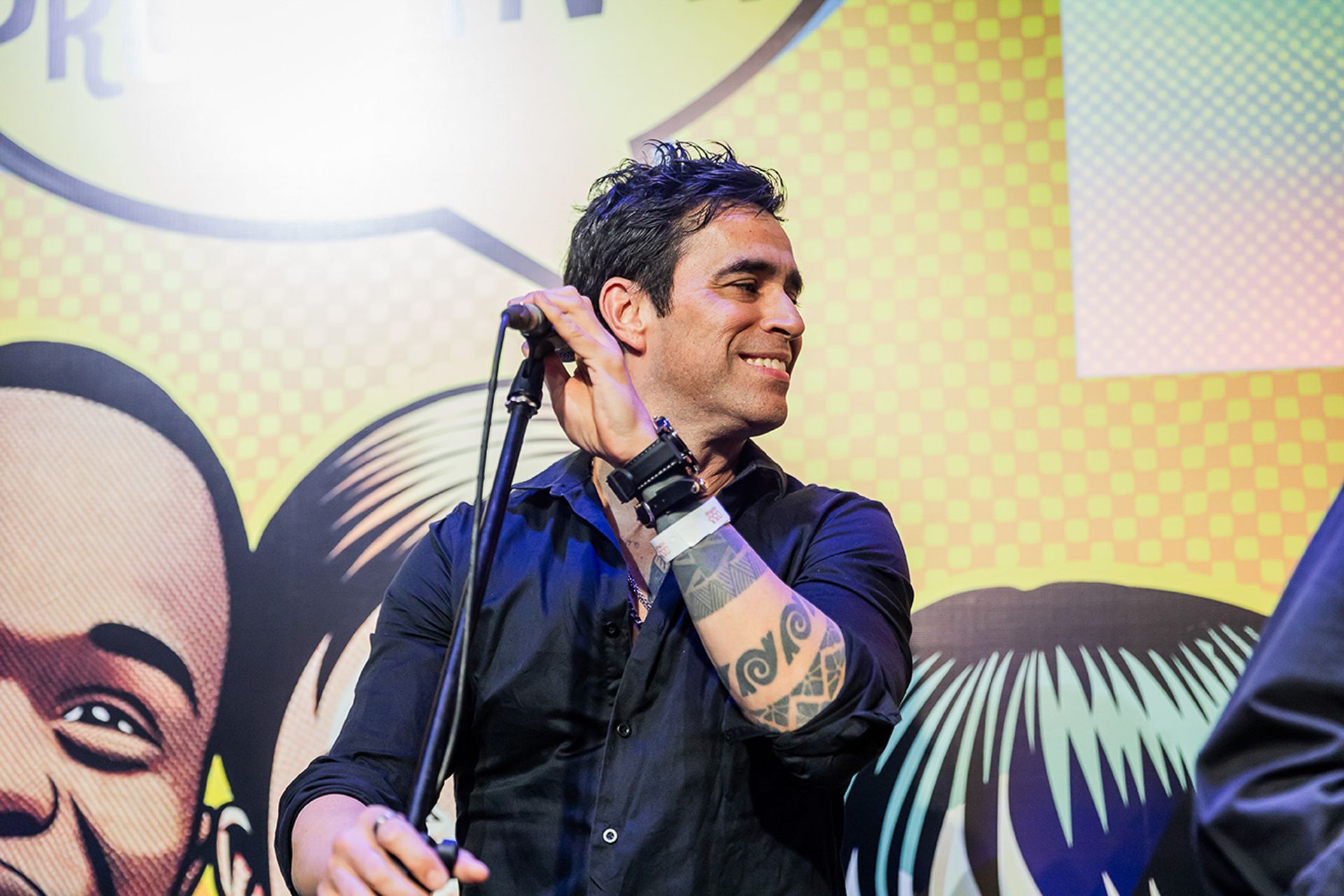 Um artista de camisa preta e braço tatuado está em pé diante de um microfone, sorrindo.